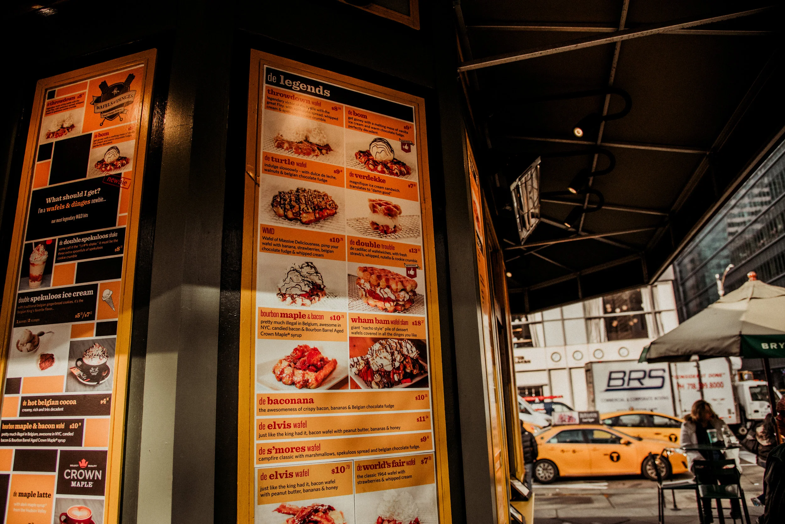 City street scene with a menu board advertising Belgian waffles outside a cafe, yellow taxis parked, a person sitting at an outdoor table, and tall buildings in the background.