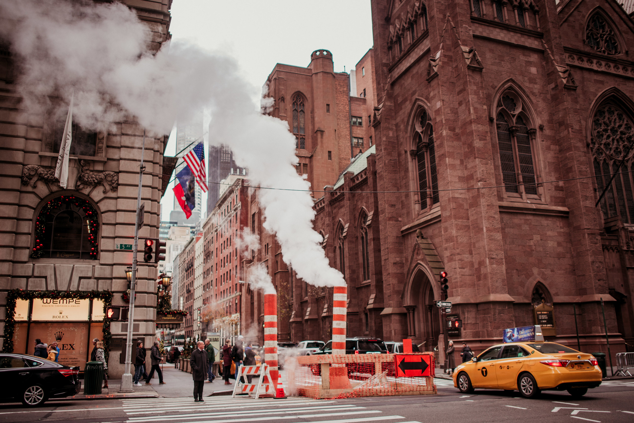 City street scene with construction barriers, steam vents, and a yellow taxi; historic brick buildings and pedestrians in the background, American flags and a decoration-adorned shop front.