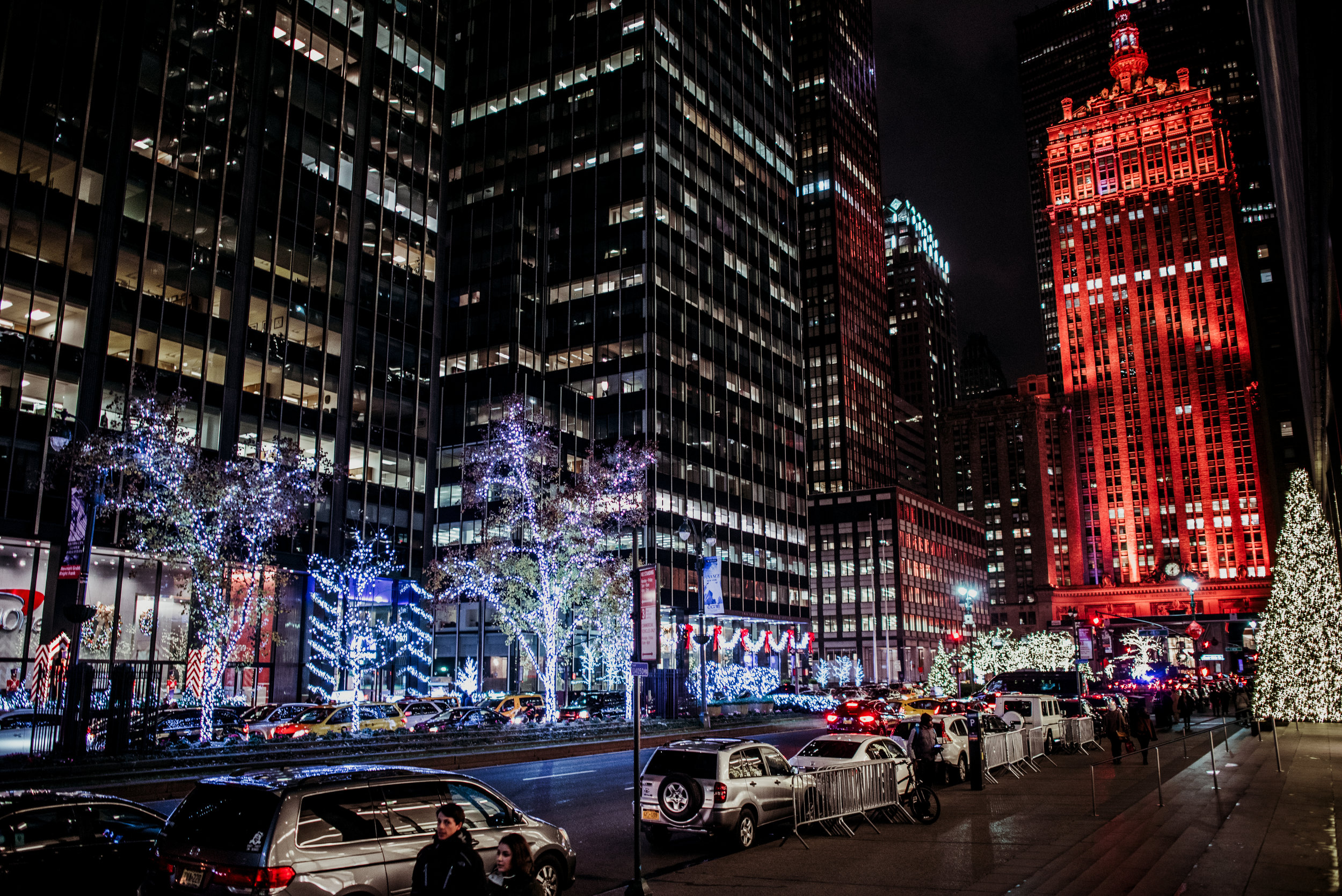 Nighttime city street with tall buildings lit up, decorated with Christmas lights, including Christmas trees and illuminated trees along the sidewalk, and cars parked along the curb.