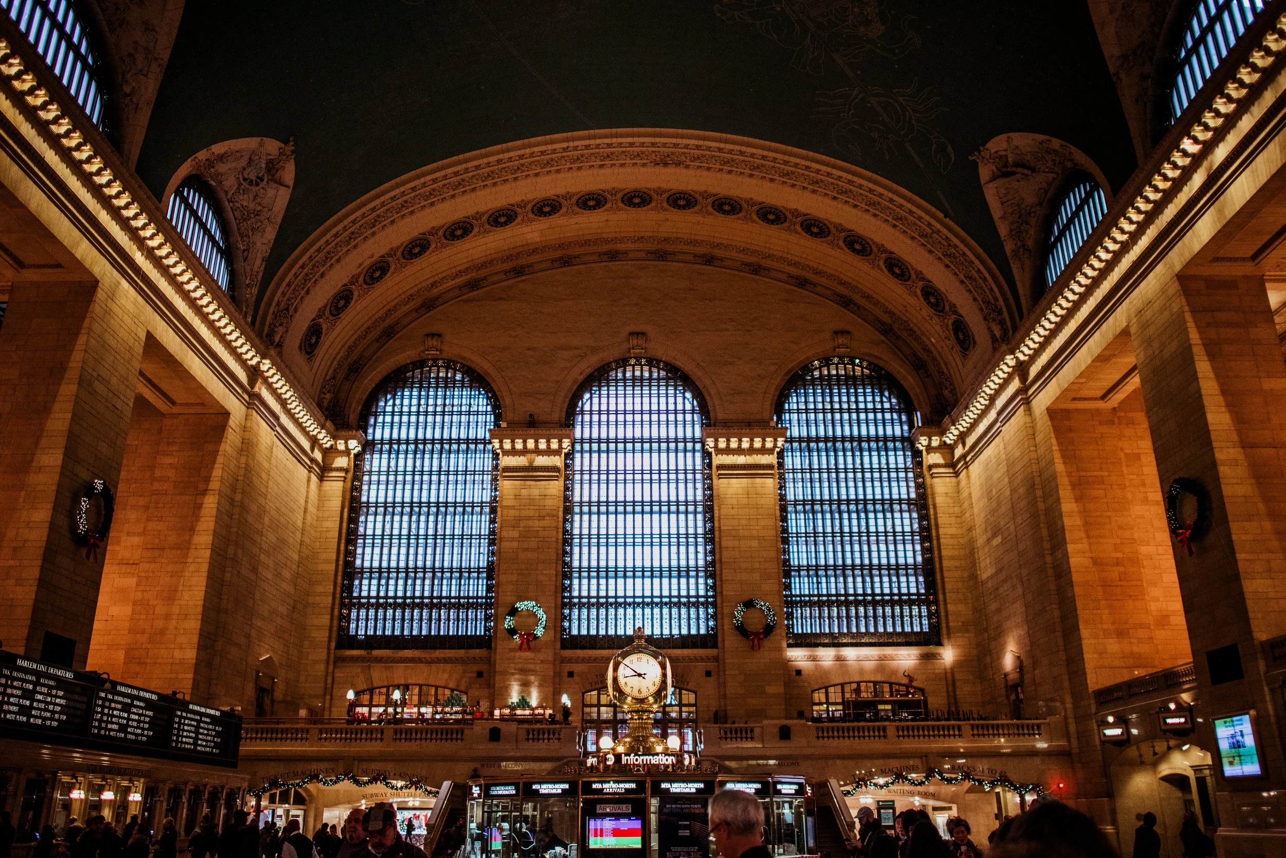 Historic train station interior with large arched windows, a clock, holiday wreaths, and Christmas decorations.