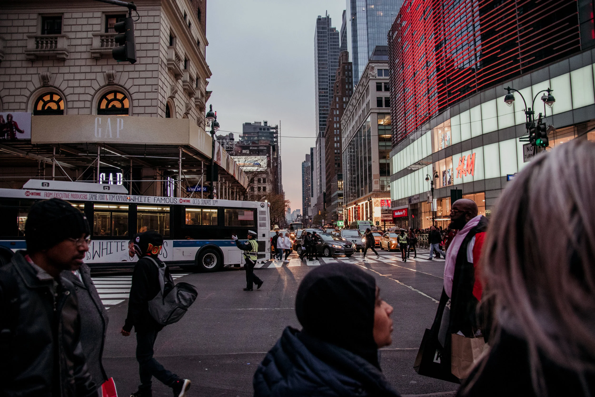 Urban city street scene with pedestrians crossing the street, taxis, and a bus, surrounded by tall buildings with store signs including H&M and GAP, during dusk.