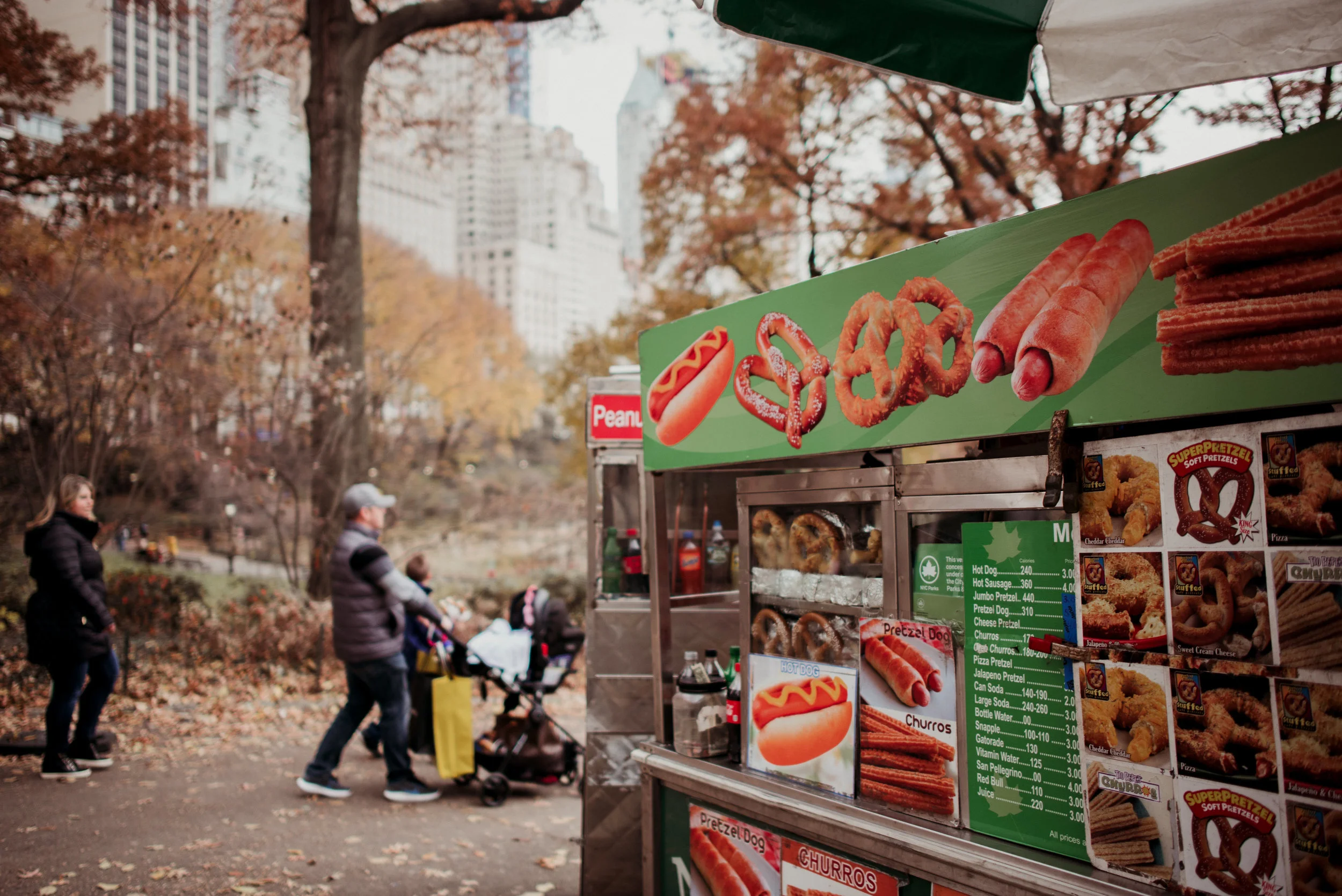 A food cart selling hot dogs, pretzels, churros, and other snacks at a park with people walking by and trees with autumn leaves in the background.