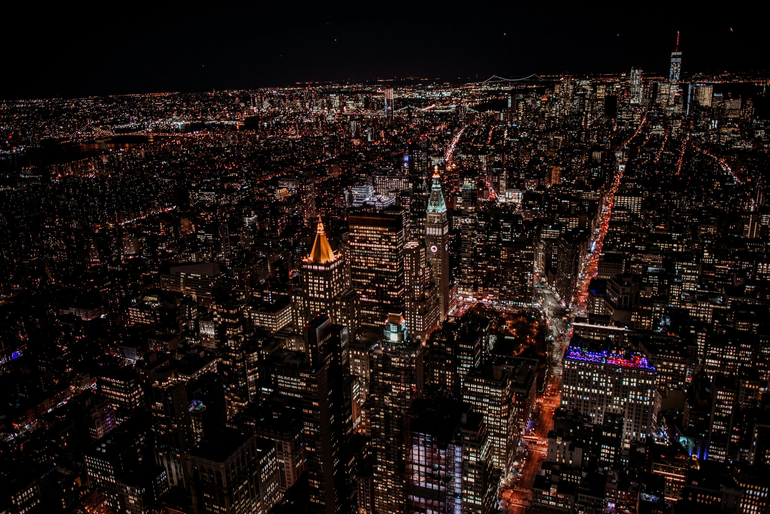 Nighttime aerial view of a city skyline with illuminated skyscrapers, including the tall clock tower, and lit streets extending into the distance.