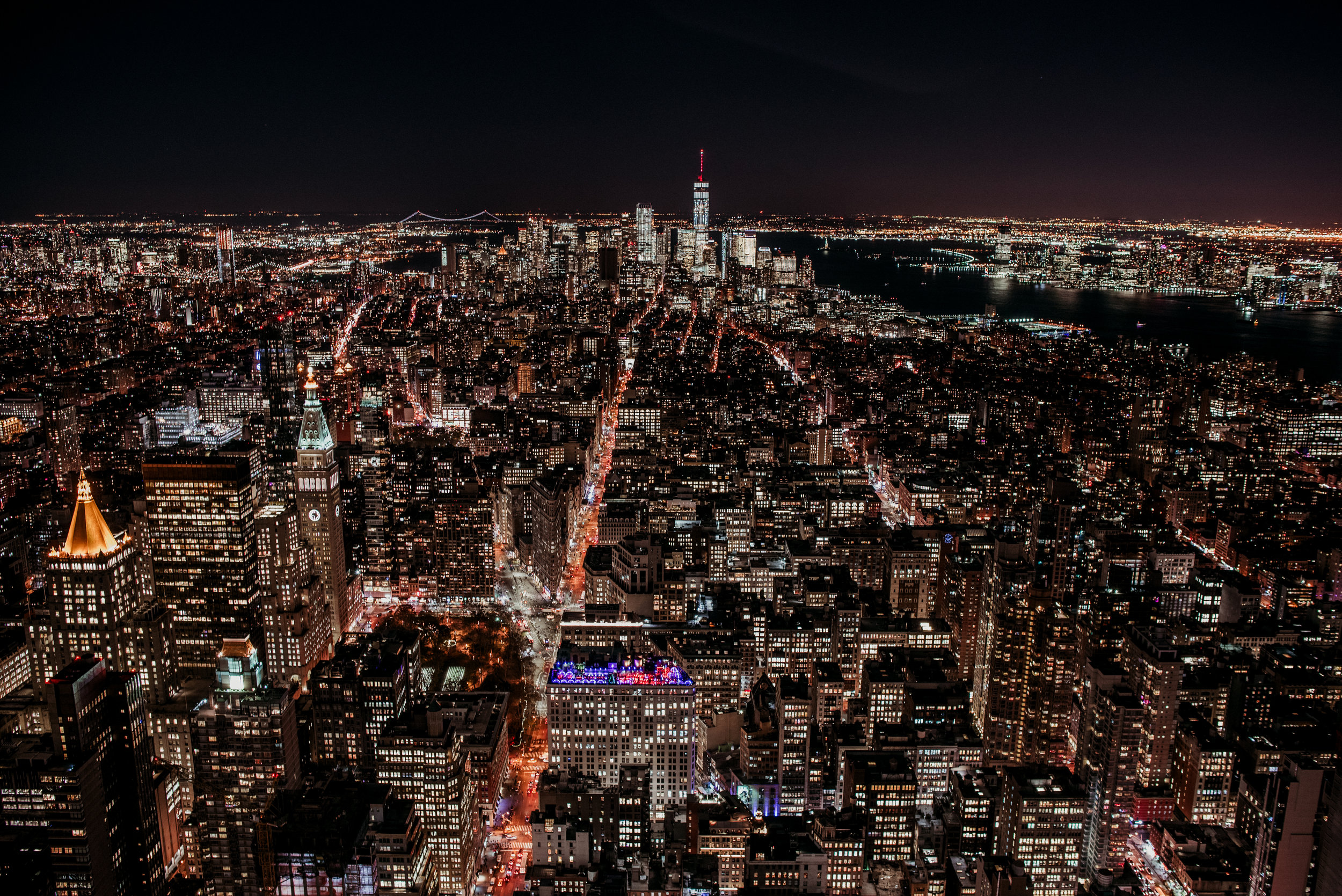 Nighttime aerial view of New York City skyline with illuminated buildings, streets, and the water in the distance.