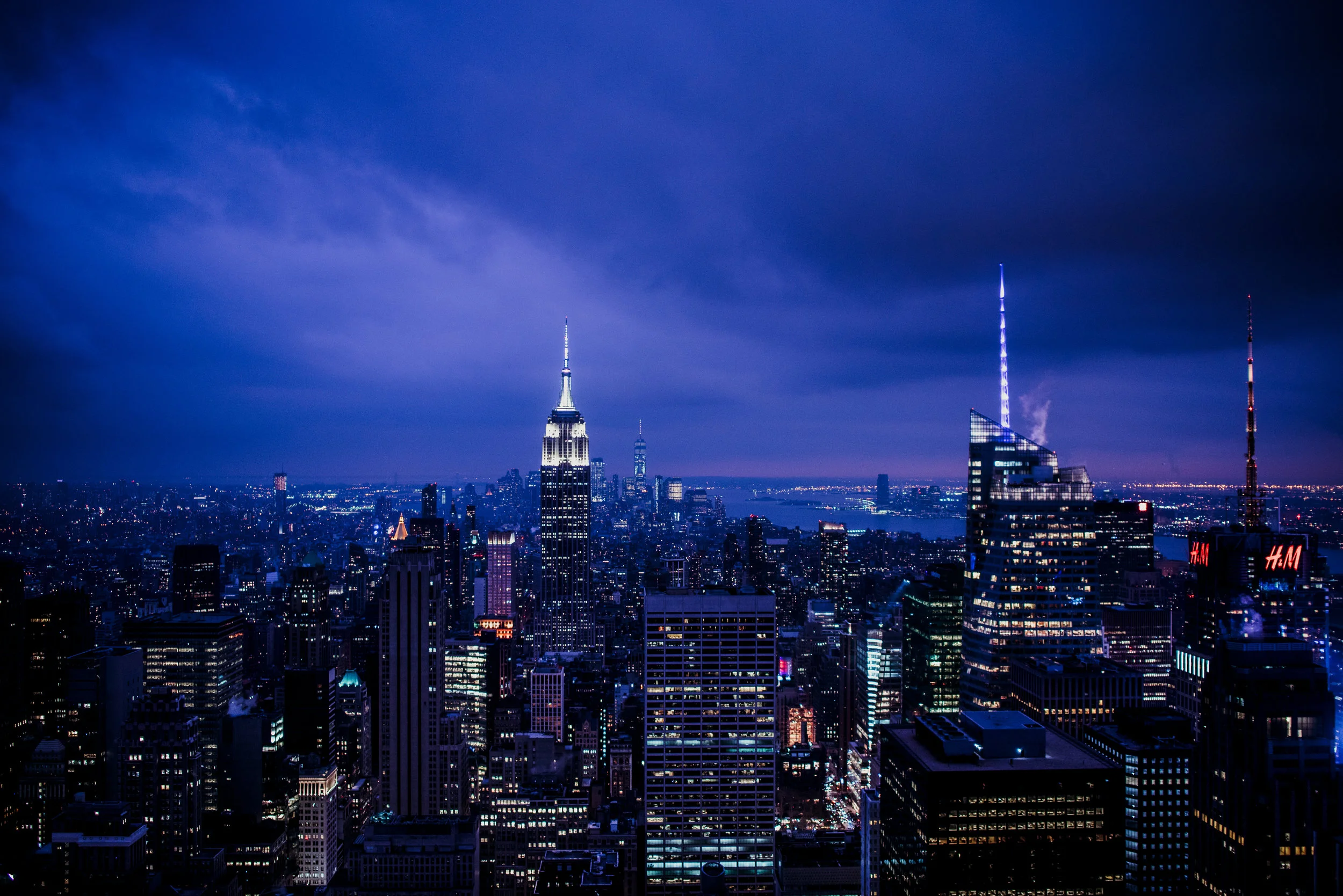 Nighttime view of New York City skyline, featuring the Empire State Building and One World Trade Center, with dark clouds overhead and city lights illuminating the scene.