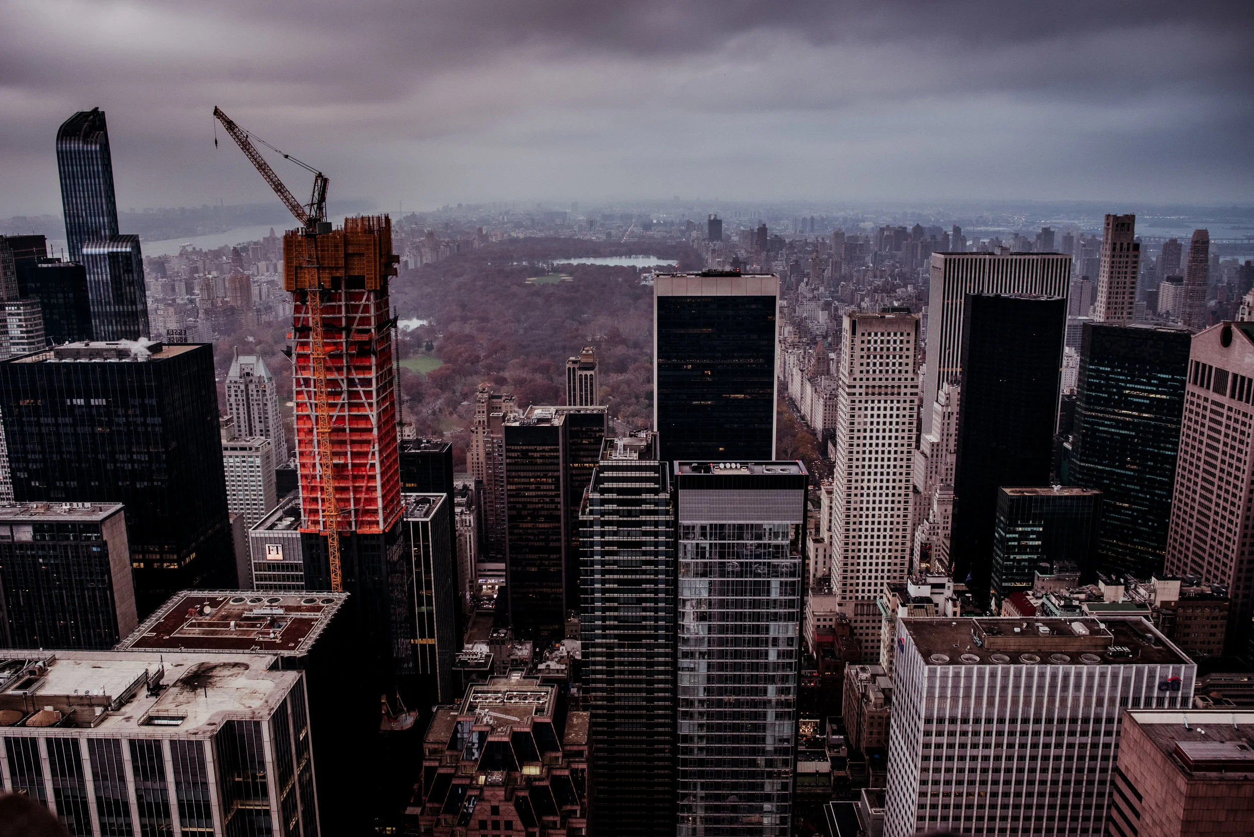 A city skyline with tall skyscrapers, a construction crane on a building under construction, and a park with trees in the background under a cloudy sky.