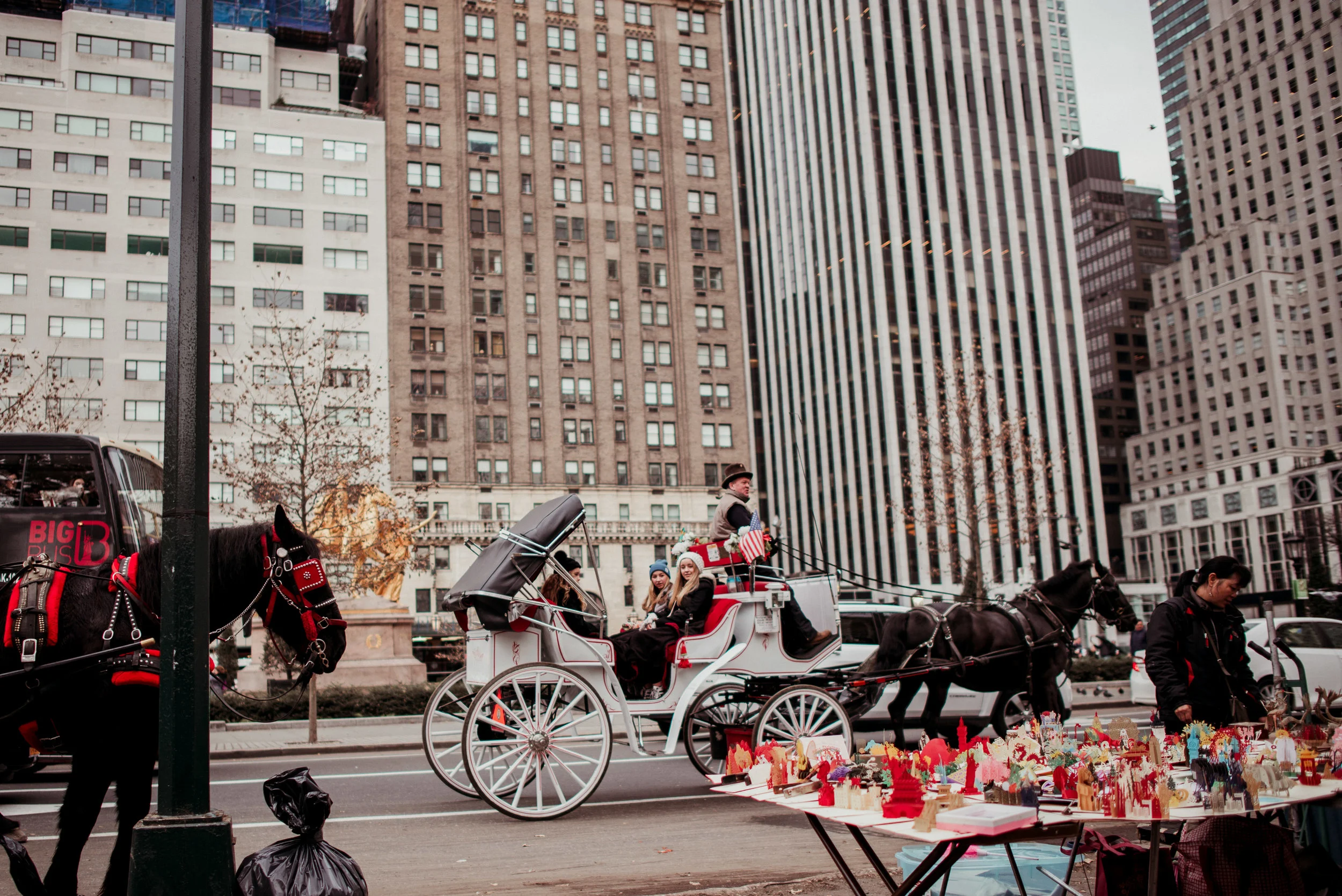 Horse-drawn carriage with people riding through city street with tall buildings in background, sidewalk vendor selling small colorful items.