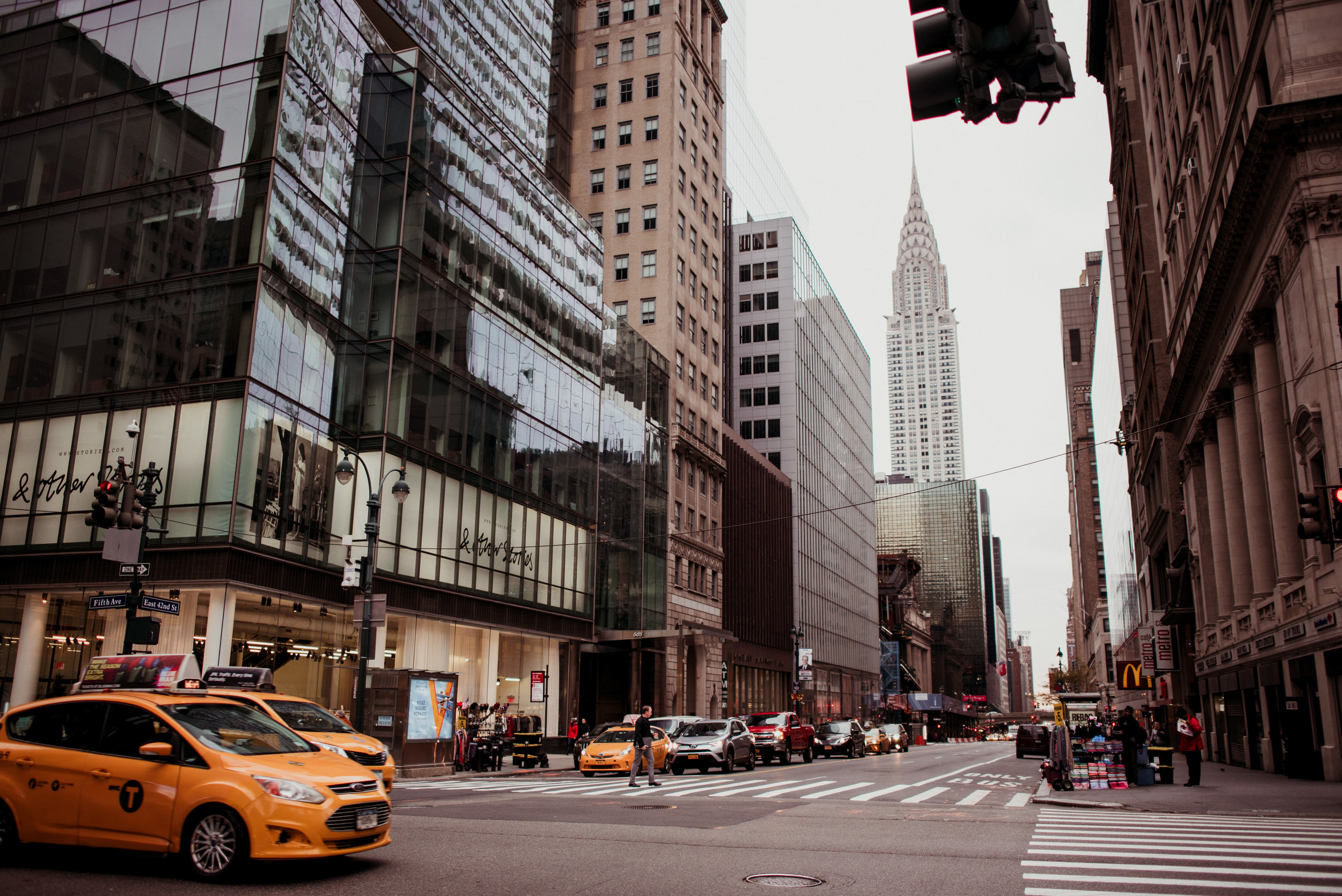 City street scene with tall buildings, yellow taxis, pedestrians, and storefronts, including a McDonald's sign, with the Chrysler Building visible in the background.