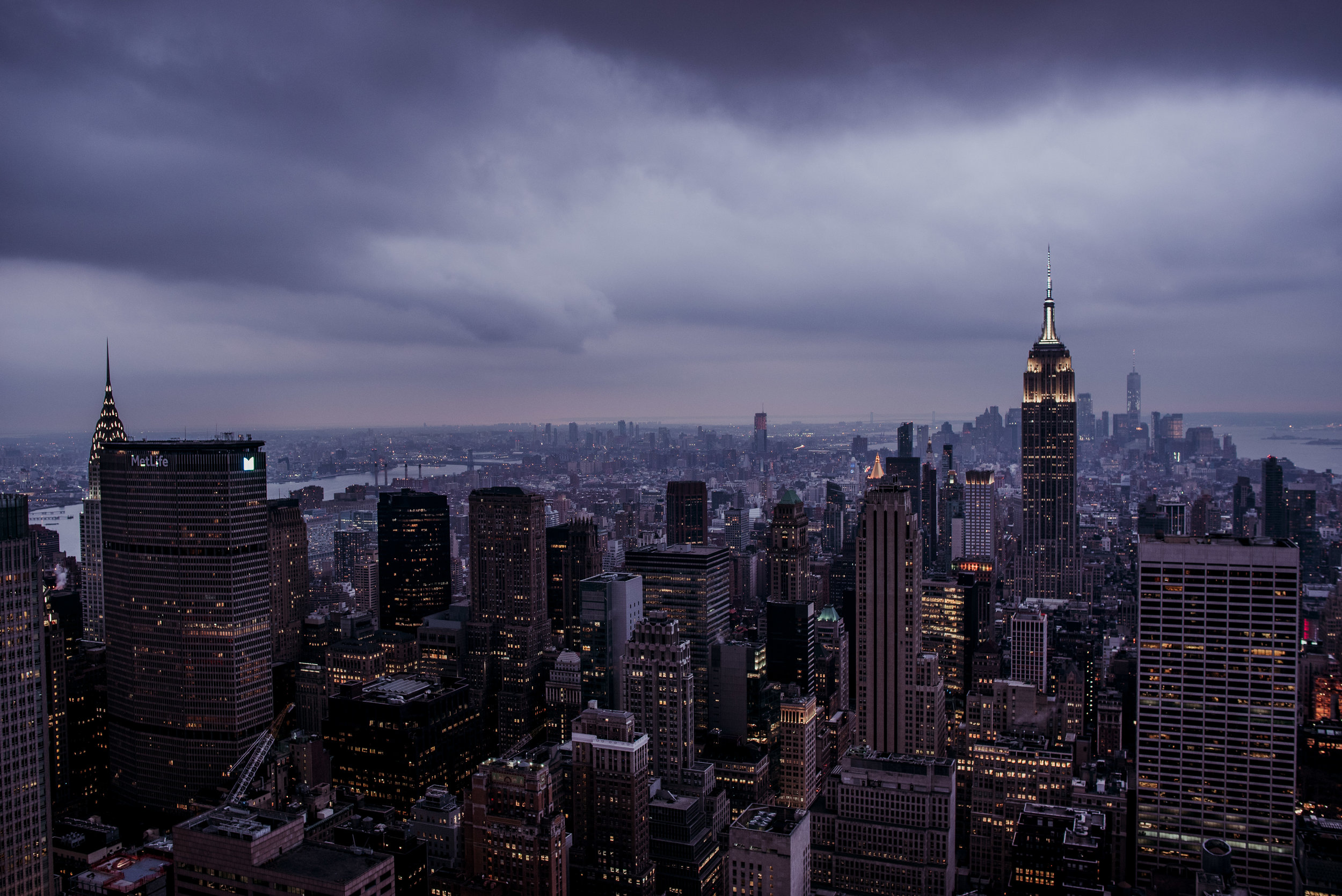 Nighttime view of New York City skyline with many tall buildings including the Empire State Building. Dark stormy clouds over the city, with lights starting to turn on.