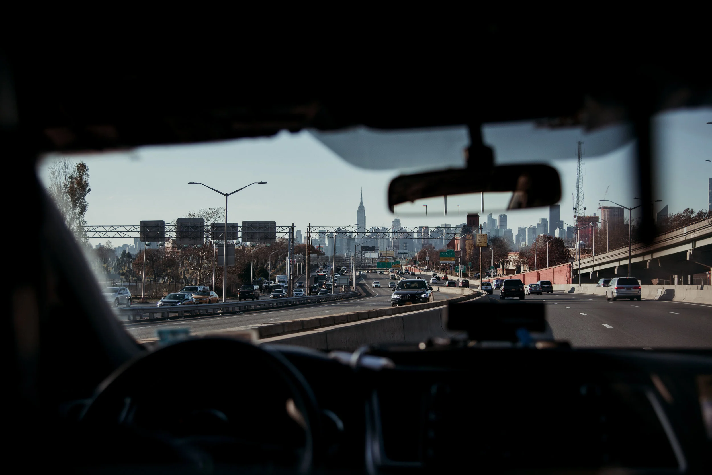 View of a busy highway leading to New York City with the skyline, including the Empire State Building, visible in the background. The photo is taken from inside a vehicle.