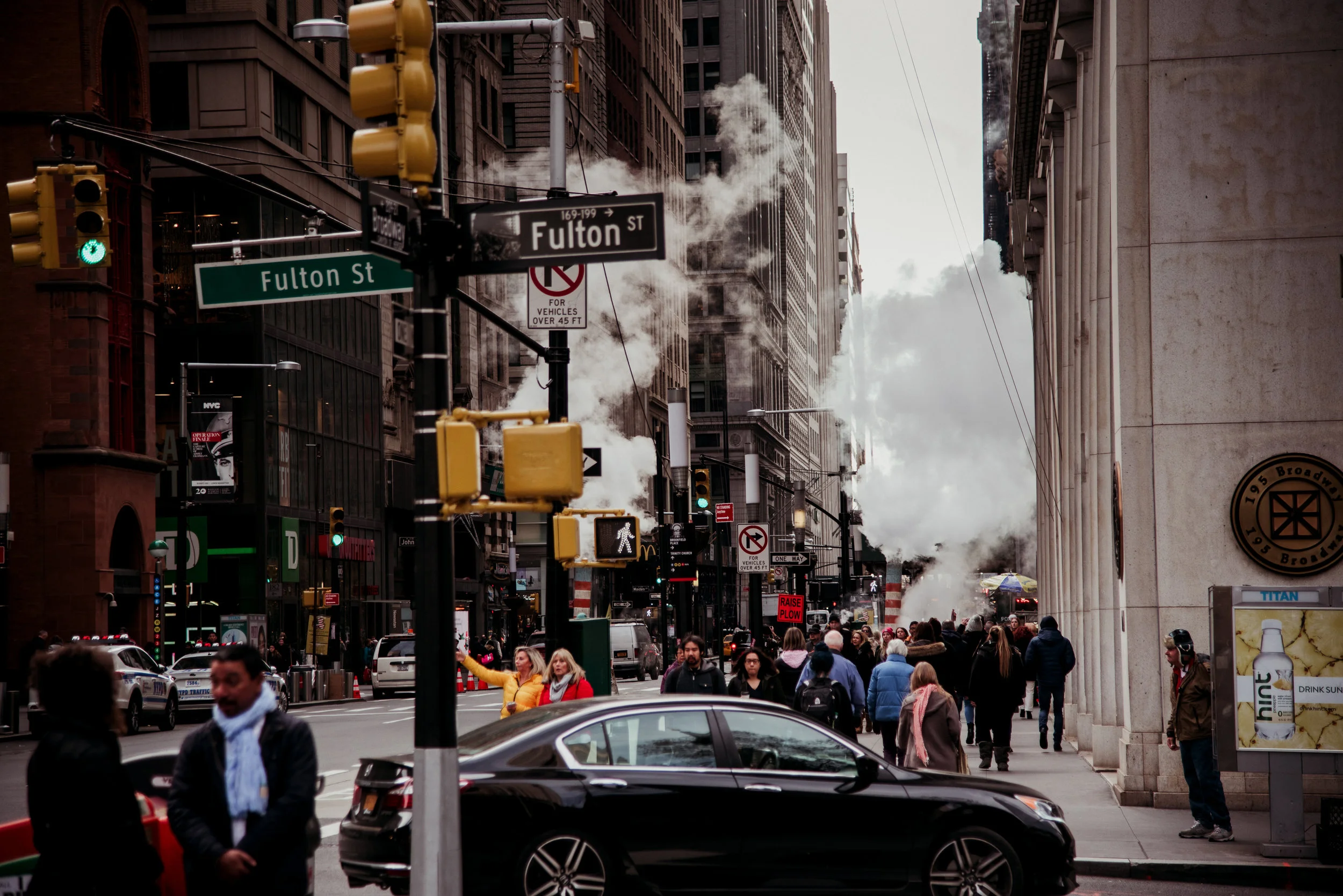 Street scene at Fulton Street in New York City with pedestrians, vehicles, street signs, and steam rising from underground pipes.