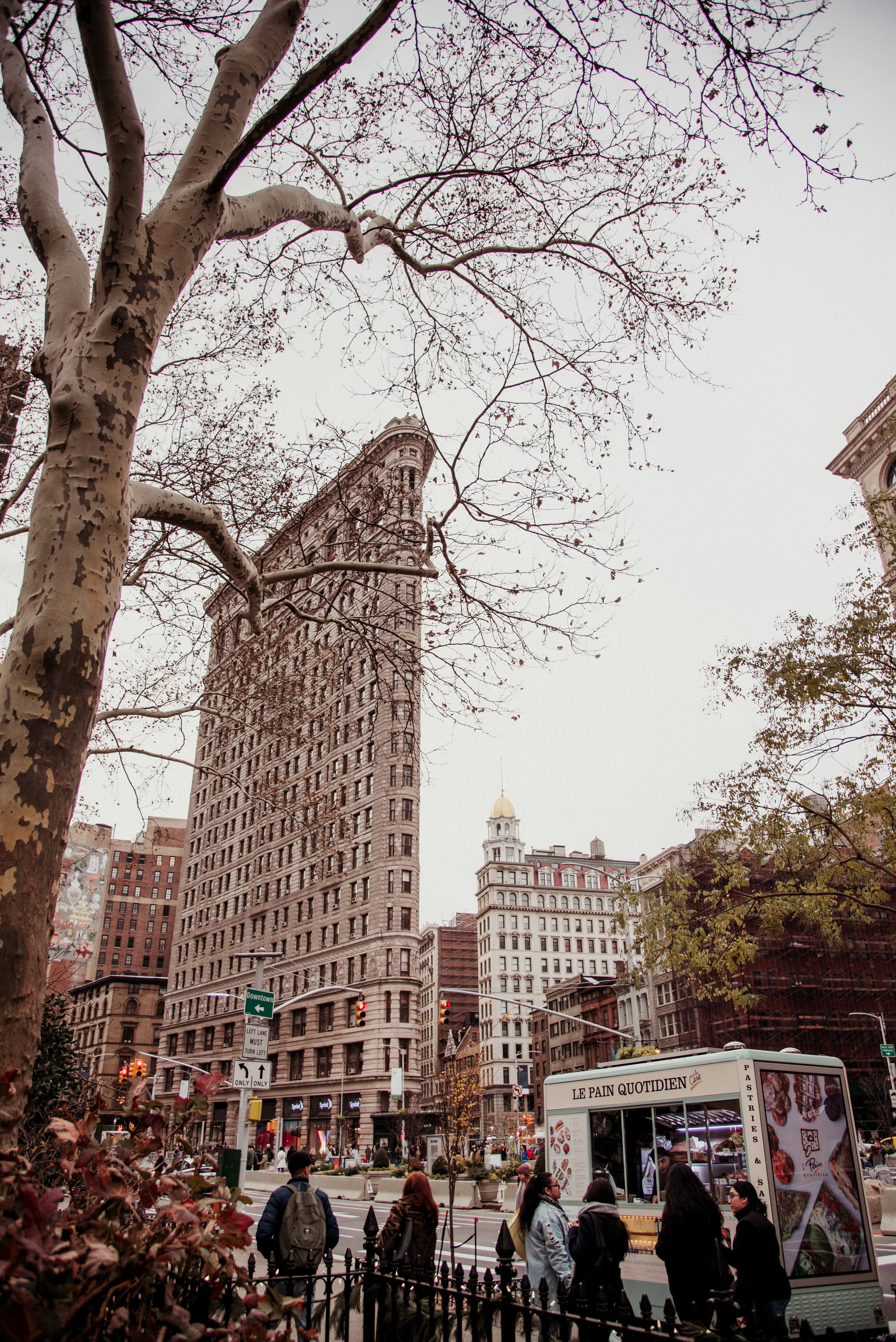 Urban street scene with tall buildings, leafless trees, a group of people, and a food truck on a cloudy day.