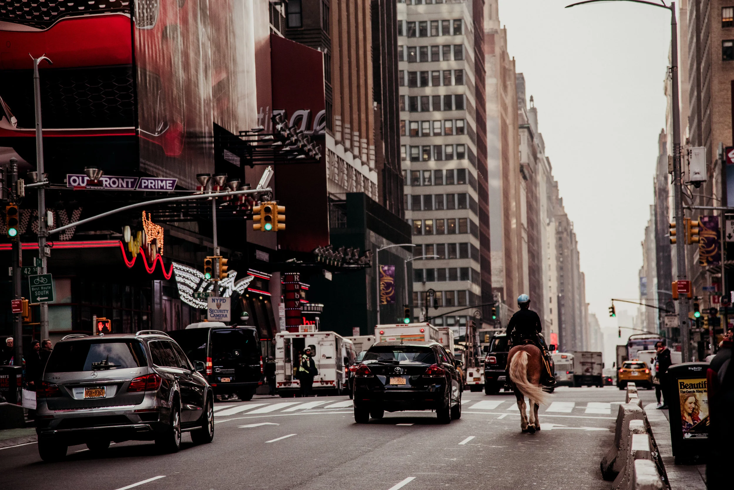 A street scene in New York City with cars, a person riding a horse, and tall buildings on both sides of the street. There are traffic lights and signs, and people standing on the sidewalk.
