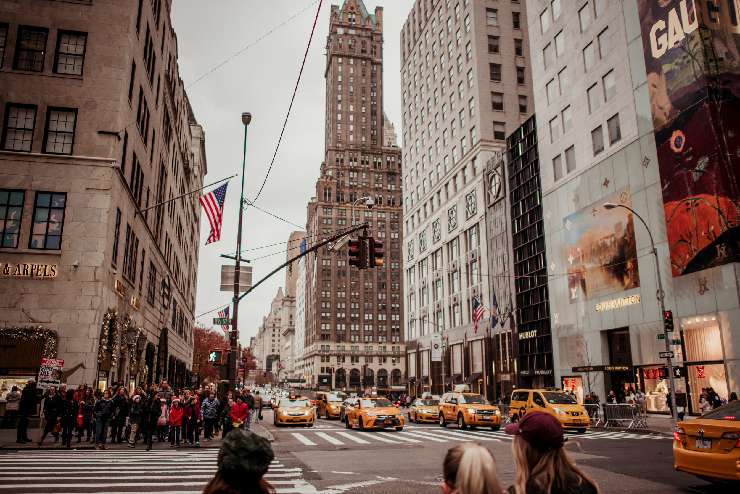 City street scene with yellow taxis, pedestrians crossing, and tall buildings including the historic Flatiron Building in the background, during overcast weather.