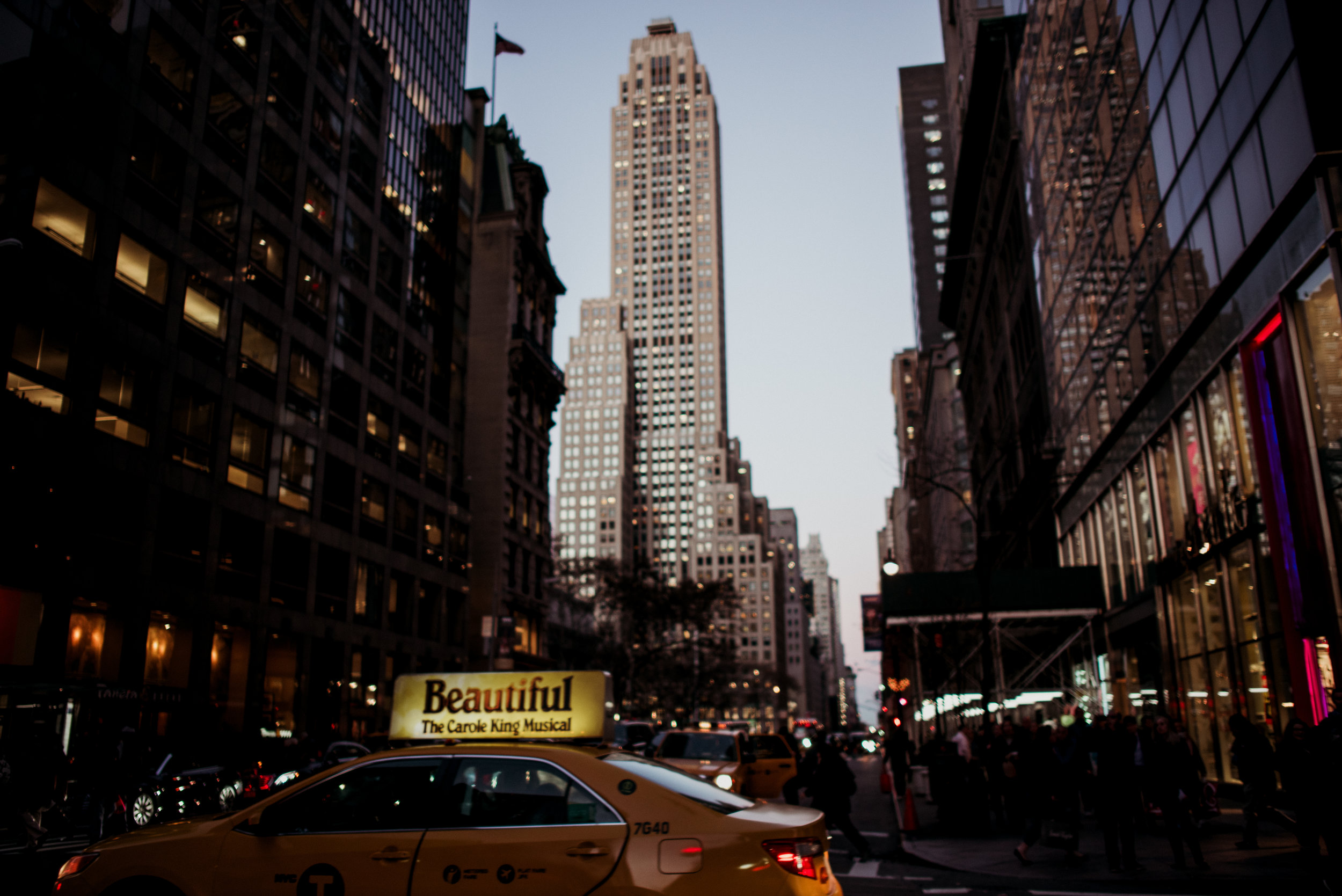 City street scene at dusk with tall skyscrapers, a yellow taxi cab in the foreground advertising a musical, and pedestrians walking along the sidewalk.