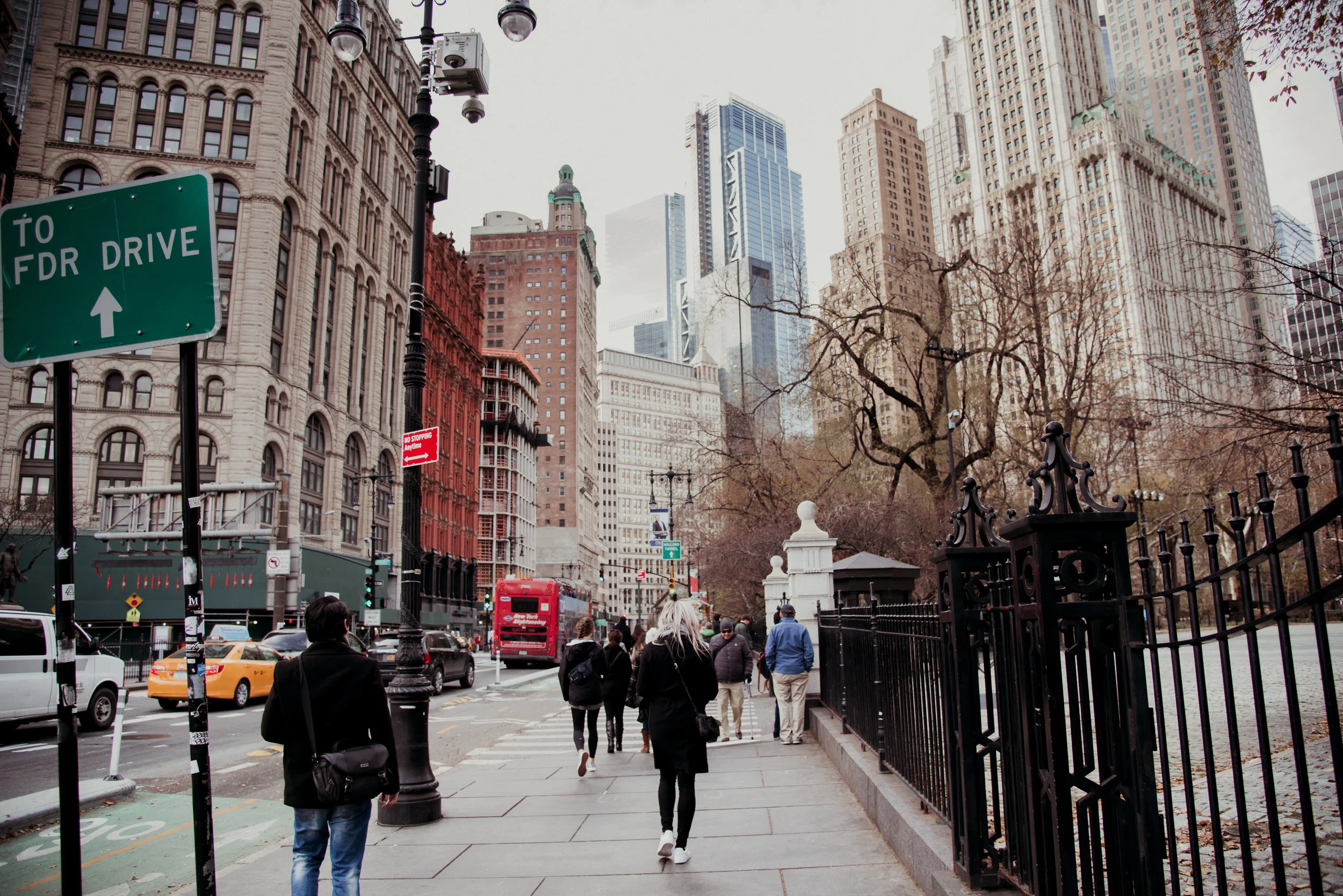 City street scene with pedestrians walking along a sidewalk next to a park with black iron fence, tall buildings and leafless trees in the background, with traffic including taxis, cars, and a double-decker bus, and street signs including a green sig