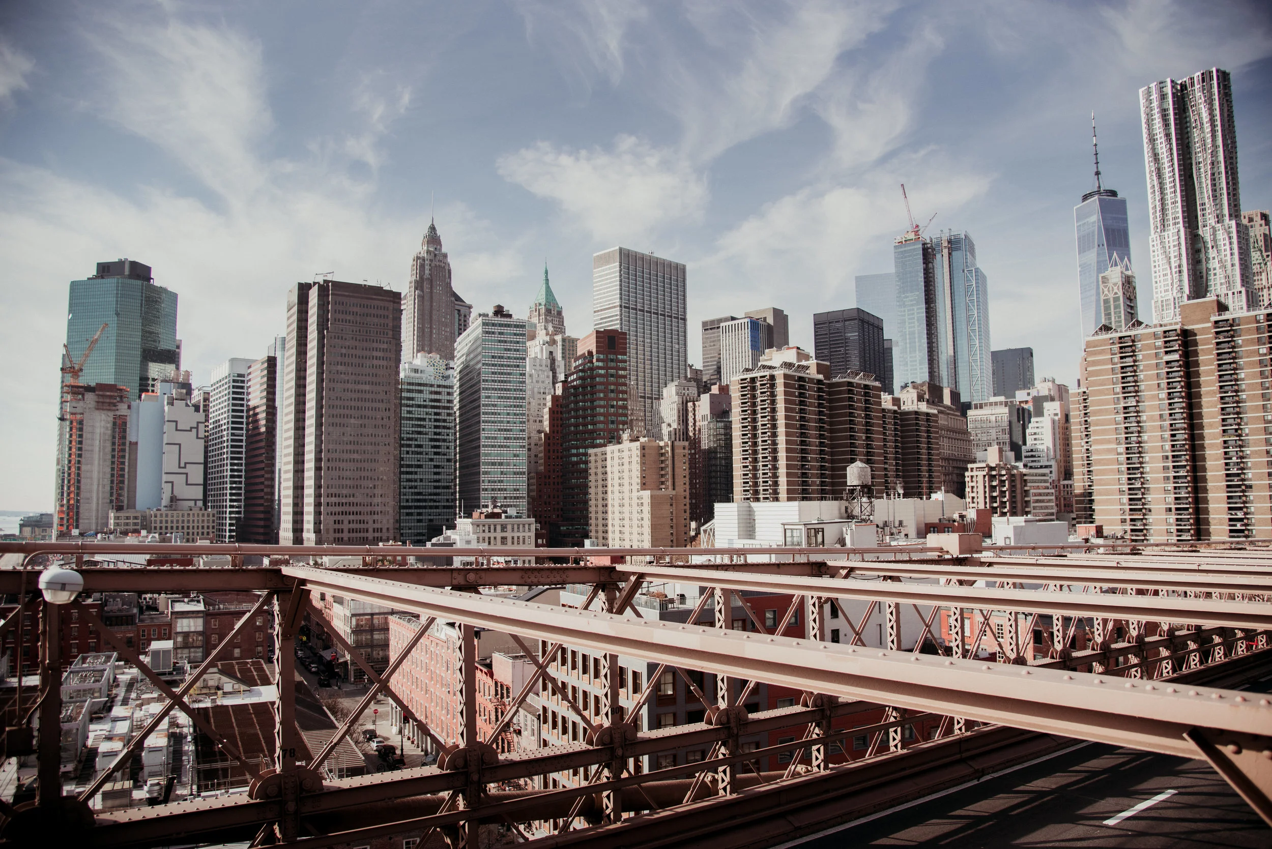 City skyline of New York City seen from a bridge, featuring tall skyscrapers under a partly cloudy sky.