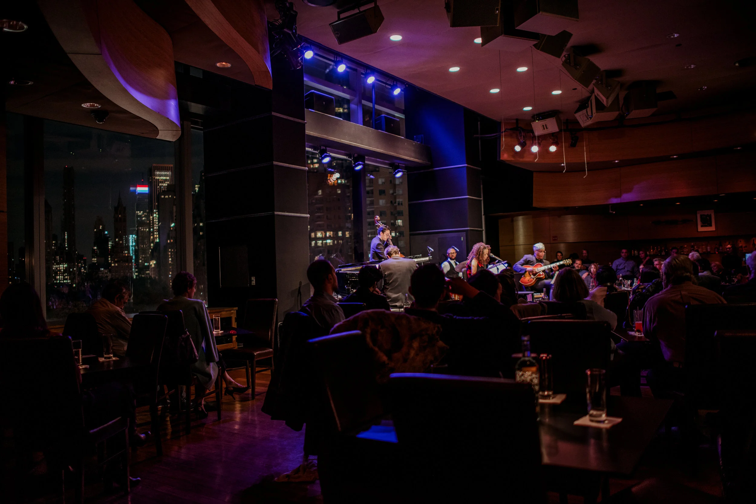 Live jazz performance at a nightclub with musicians playing piano, bass, drums, and guitar, audience seated watching under dim, colorful lighting, city skyline visible through windows.