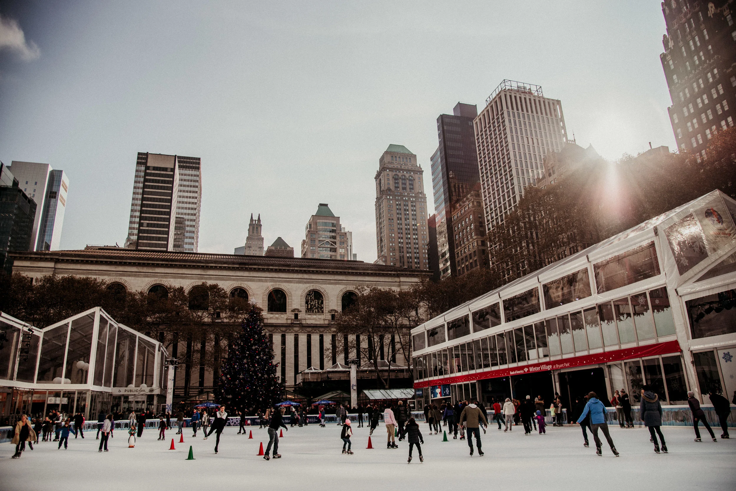 People skating on an outdoor ice rink in a city park with tall buildings, a Christmas tree, and a festive pavilion in the background during daytime.