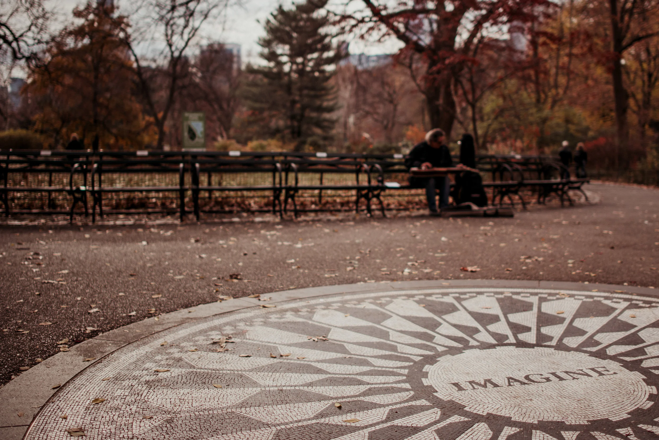 A park with a circular mosaic with the word 'IMAGINE' in the center, surrounded by autumn trees and benches. Two people are sitting on a bench, and other visitors are walking in the background.