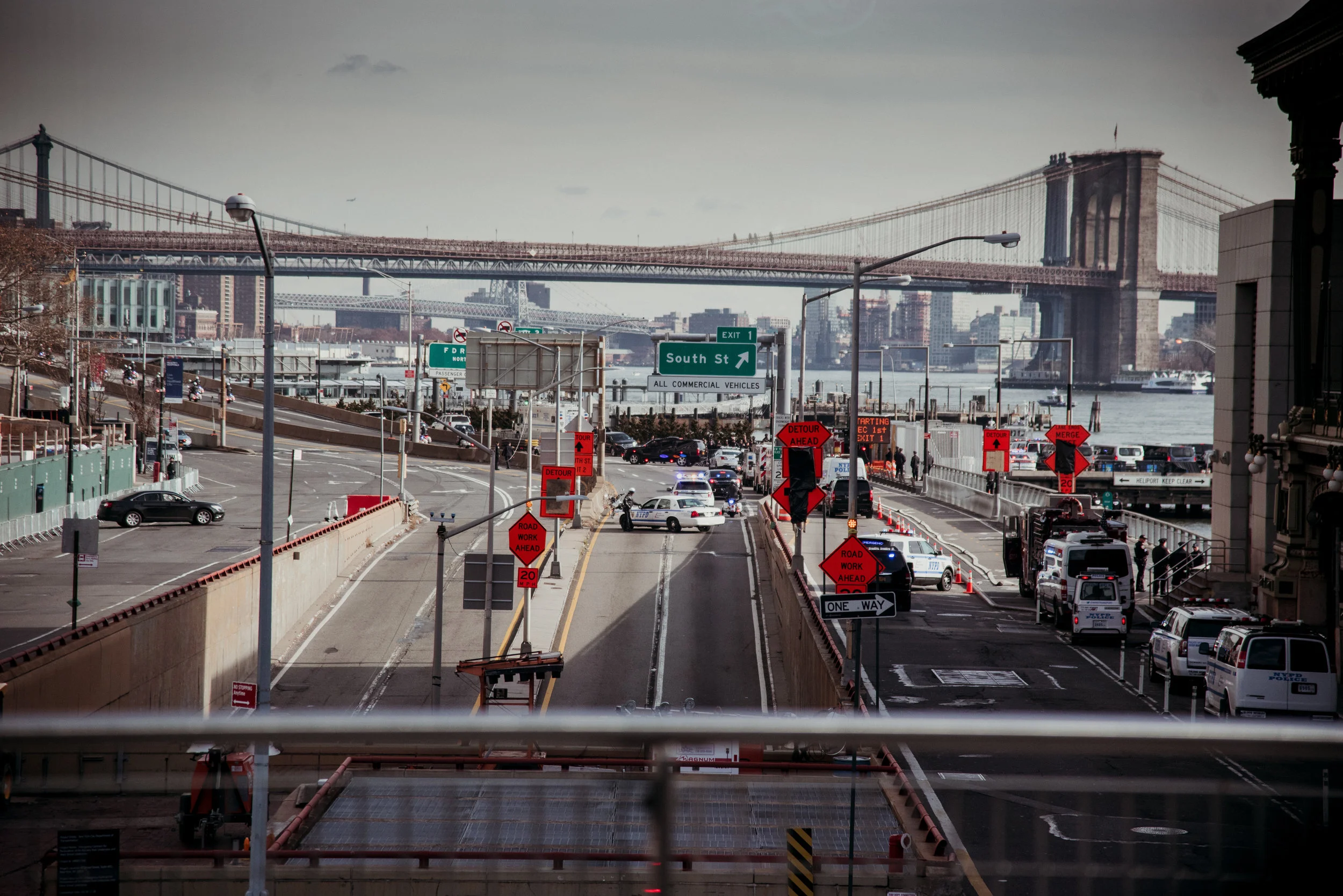 View of a busy street near a river with bridge in the background, cars, police cars, and construction signs indicating road work and detours.