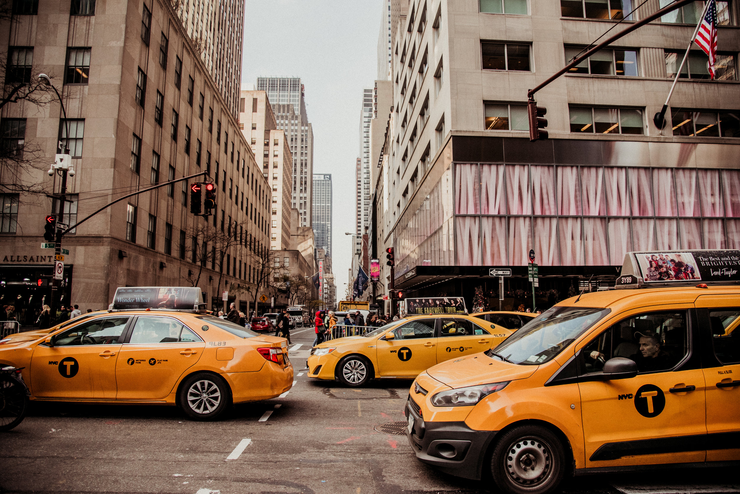 City street scene with yellow NYC taxis stopped at traffic lights, tall buildings, and pedestrians on the sidewalks.