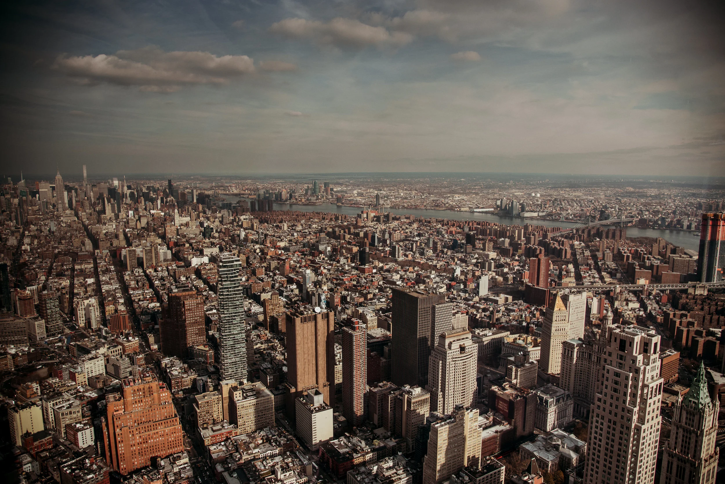 Aerial view of New York City skyline with tall skyscrapers, the East River, and bridges under a cloudy sky.