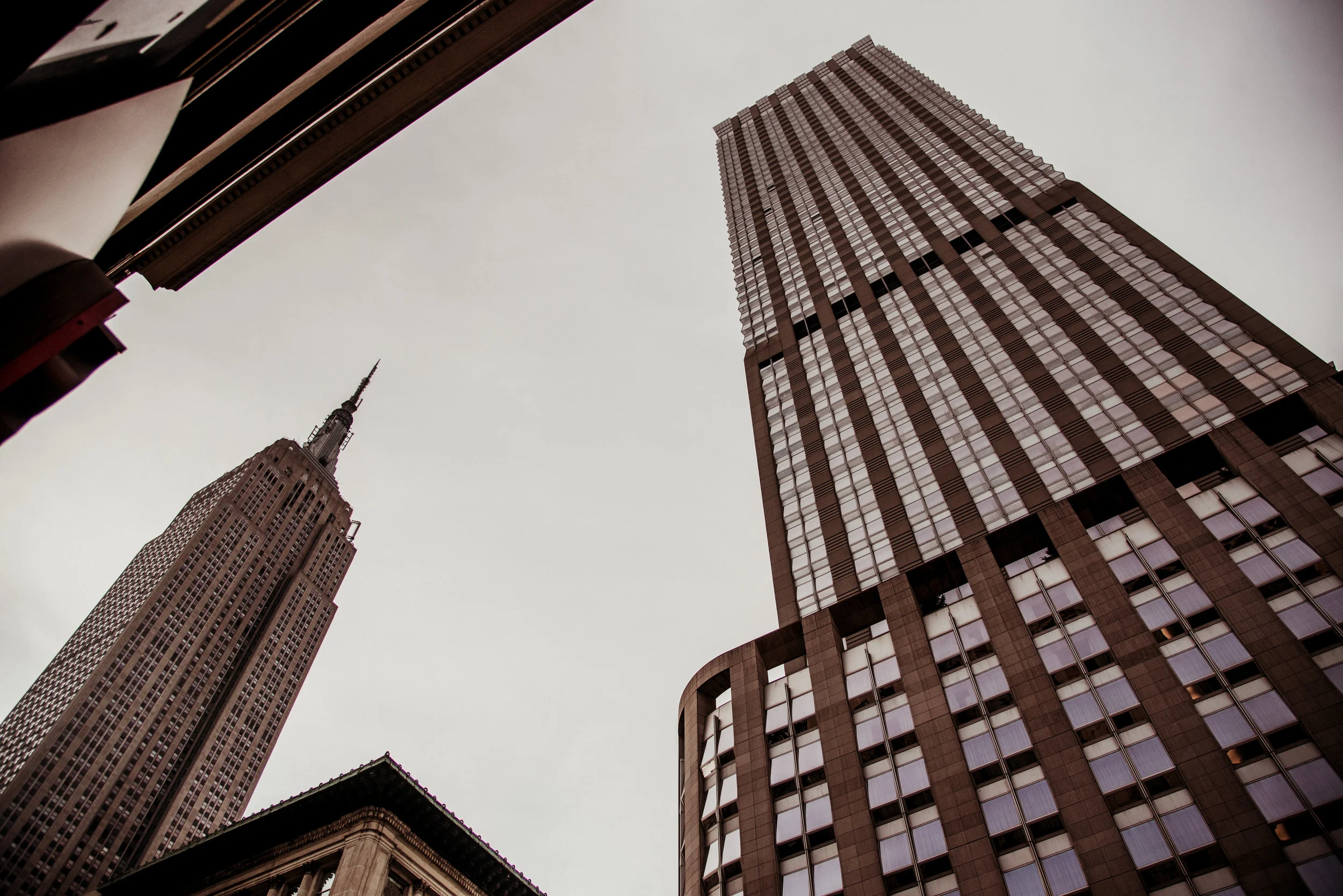 Looking up at tall skyscrapers in New York City, including the Empire State Building and Bank of America Tower, with a cloudy sky overhead.