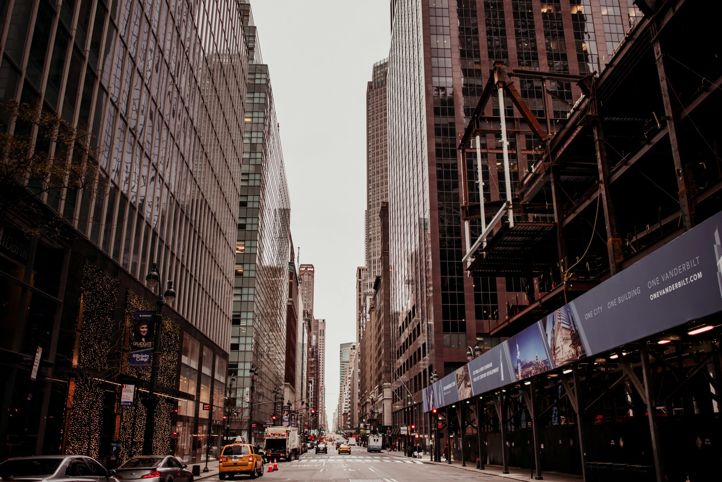 A busy city street flanked by tall glass skyscrapers, with a few cars and taxis, some pedestrians, and digital billboards on the right side.