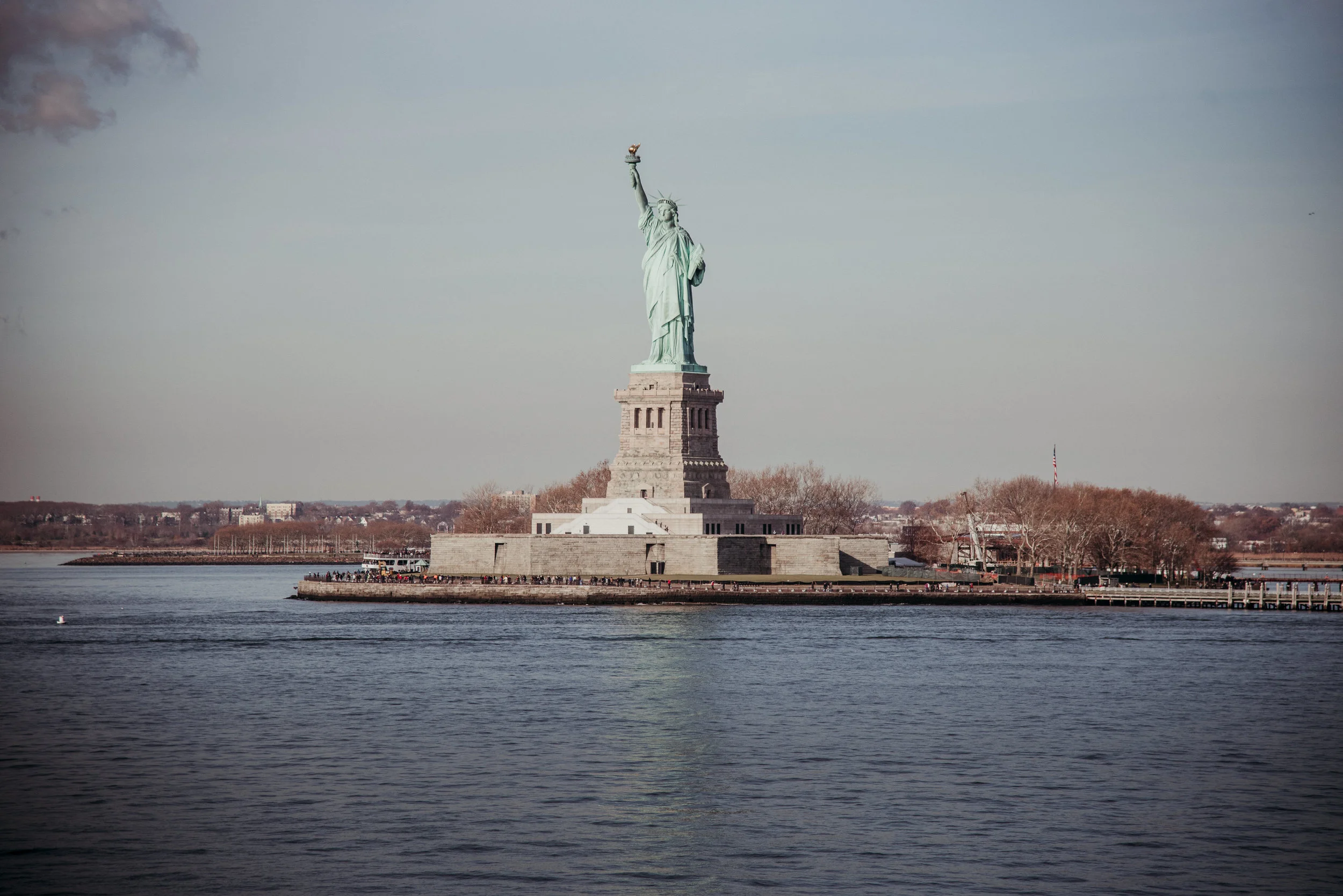 Statue of Liberty on Liberty Island in New York Harbor, with water in the foreground and a cityscape and trees in the distance.