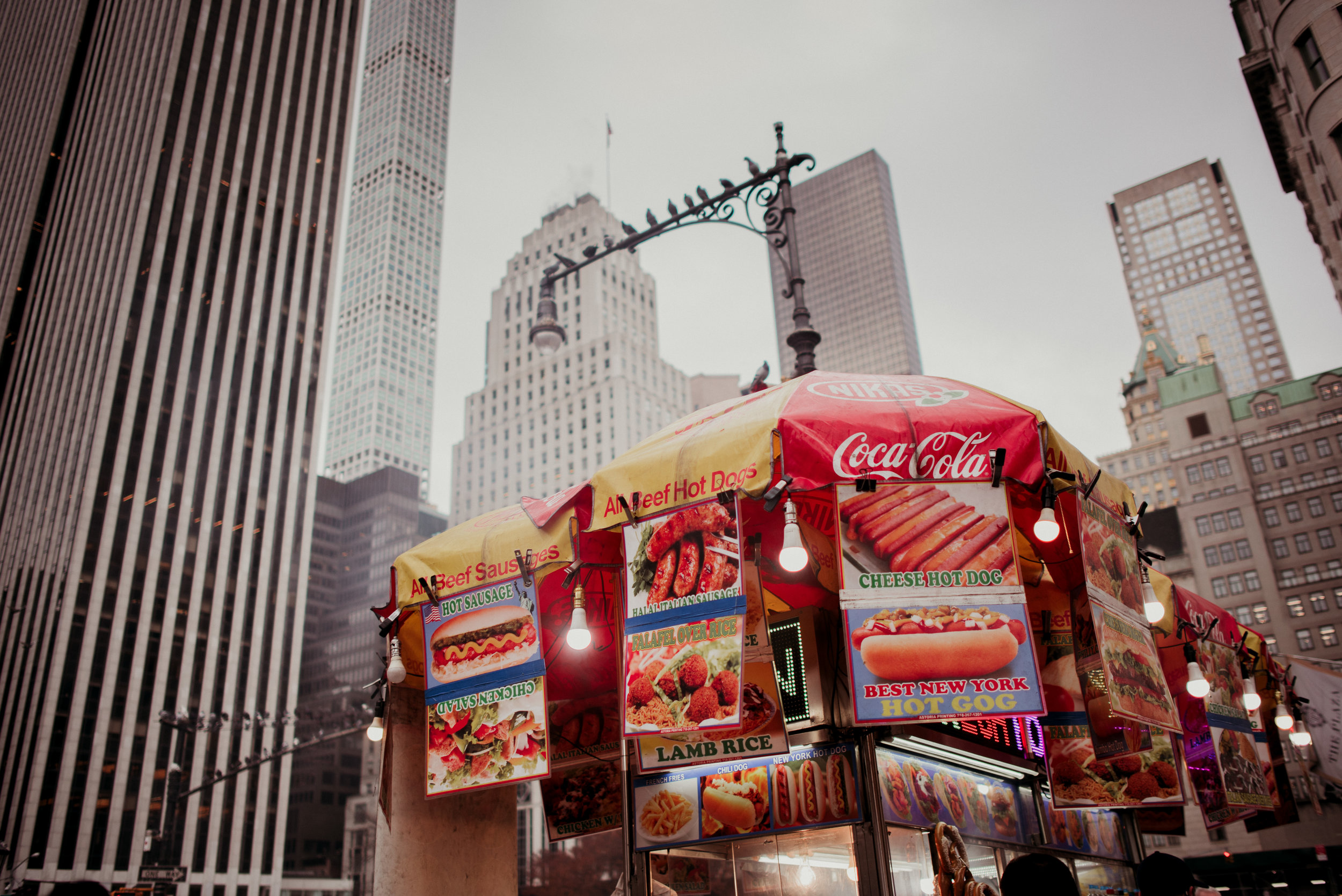 A street food cart with colorful signs advertising hot dogs, cheeseburgers, lamb rice, and sausages, set against a backdrop of tall skyscrapers in an urban cityscape