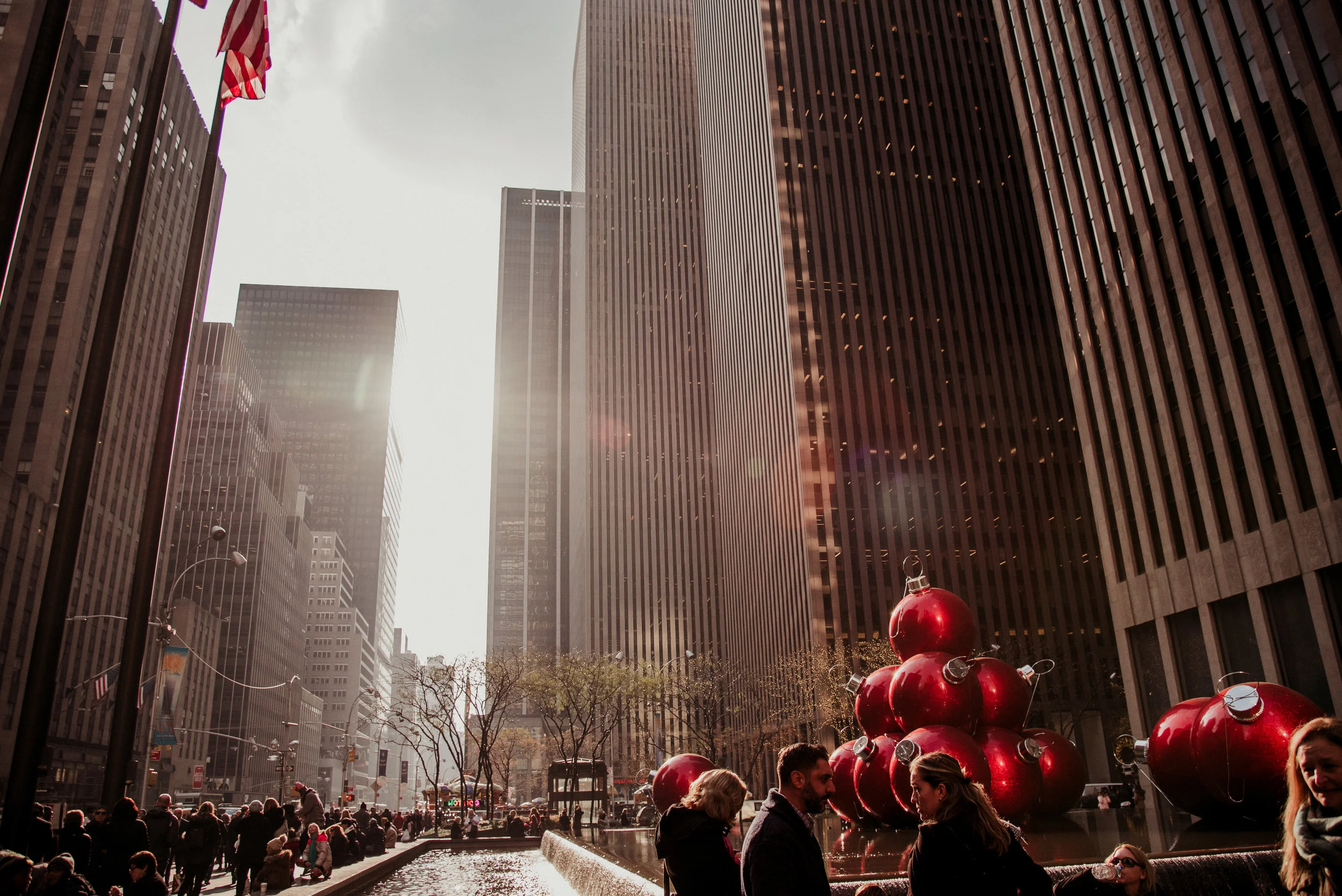 Cityscape with tall buildings, a fountain, and large red Christmas ornaments in a public space, with people walking and socializing.