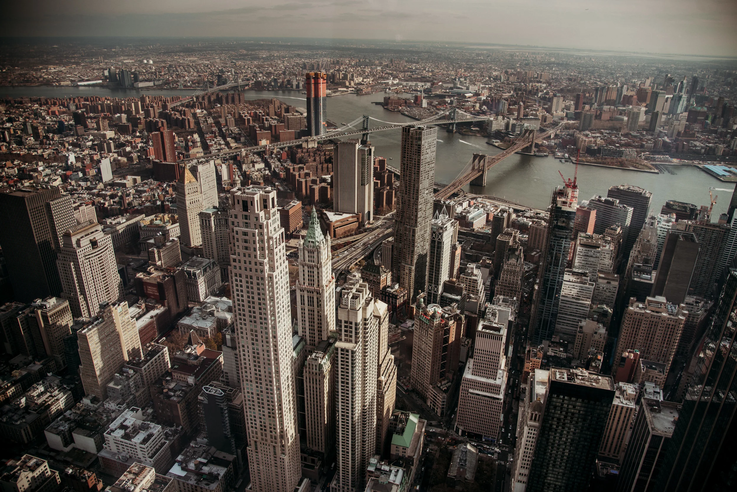 Aerial view of New York City with skyscrapers, bridges over the river, and the urban landscape under overcast sky.