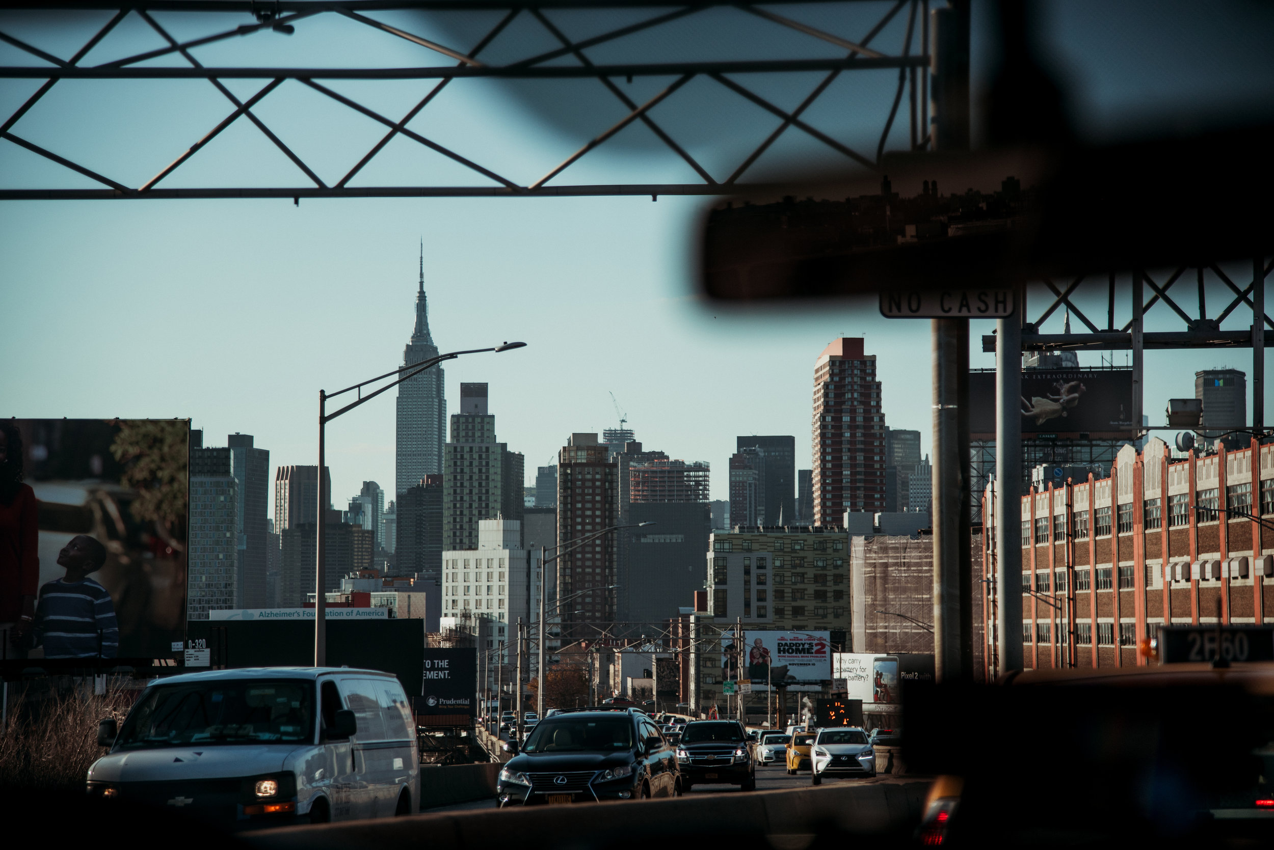 City skyline view of Manhattan with the Empire State Building in the background, seen through a car dashboard and windshield with traffic on the street in the foreground.
