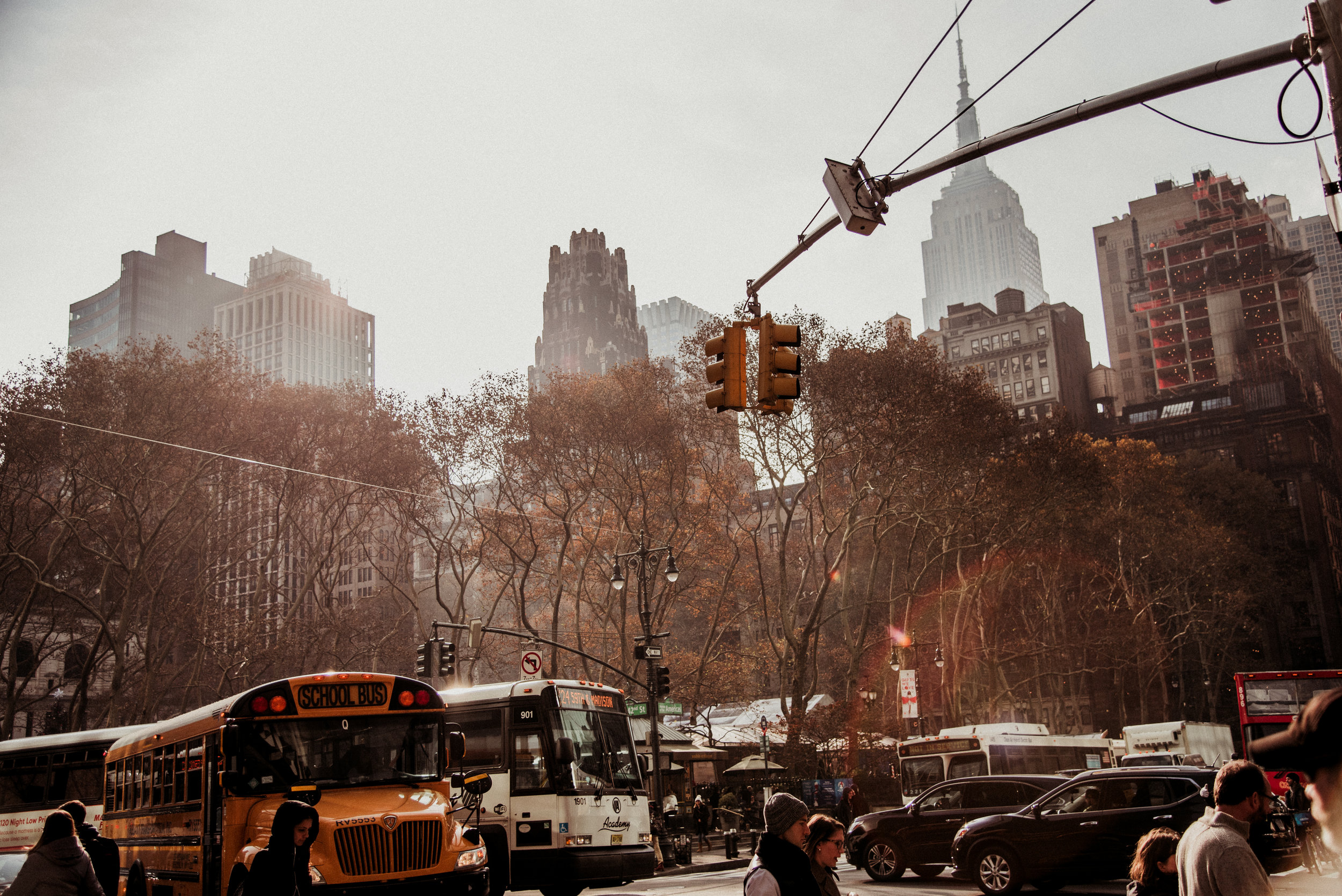 Street scene in a city with buses, cars, pedestrians, traffic lights, and tall buildings in the background, including the Empire State Building.