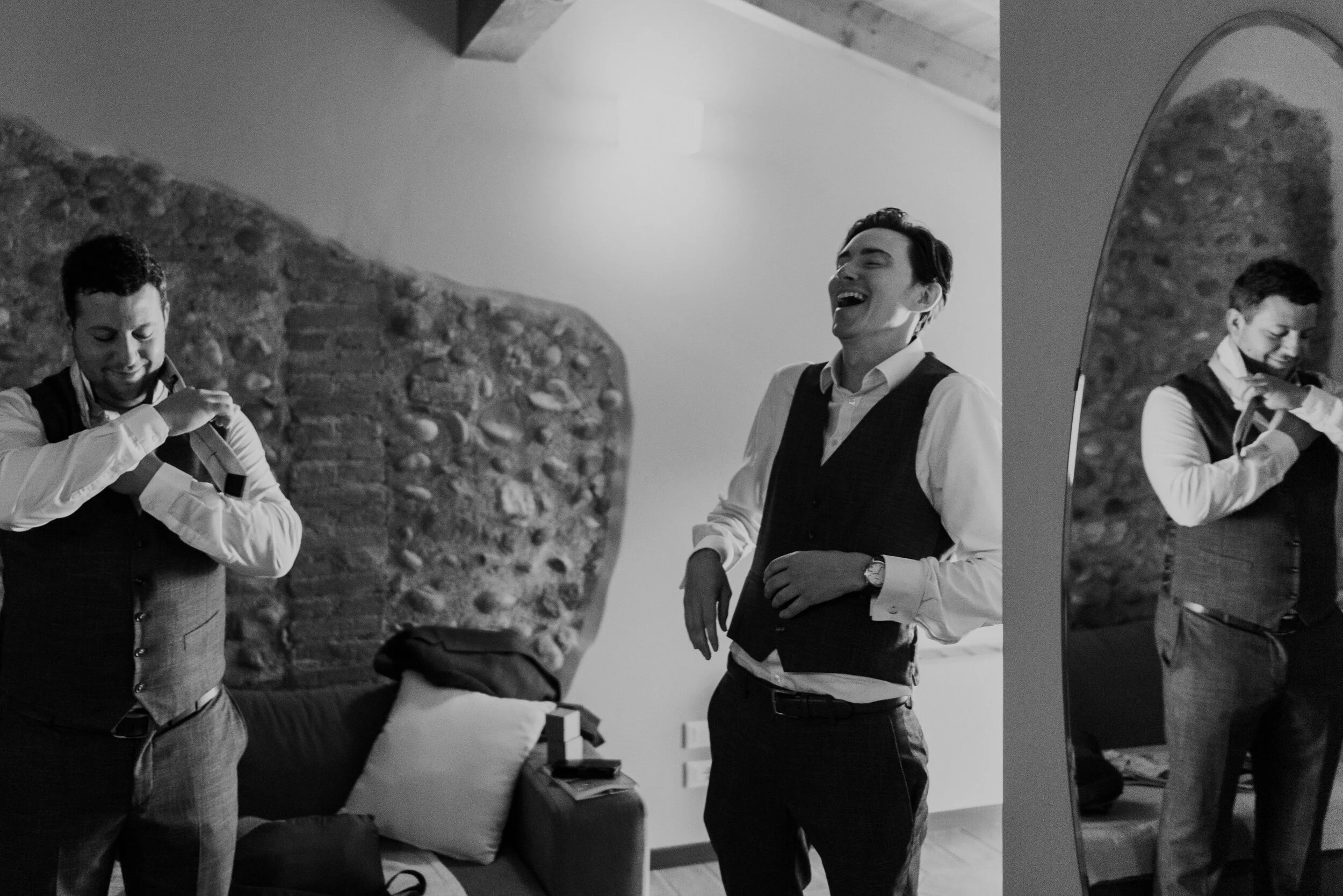 Two men dressed in formal attire, one fixing his vest and the other laughing, in a room with a brick wall and a mirror reflecting the scene.