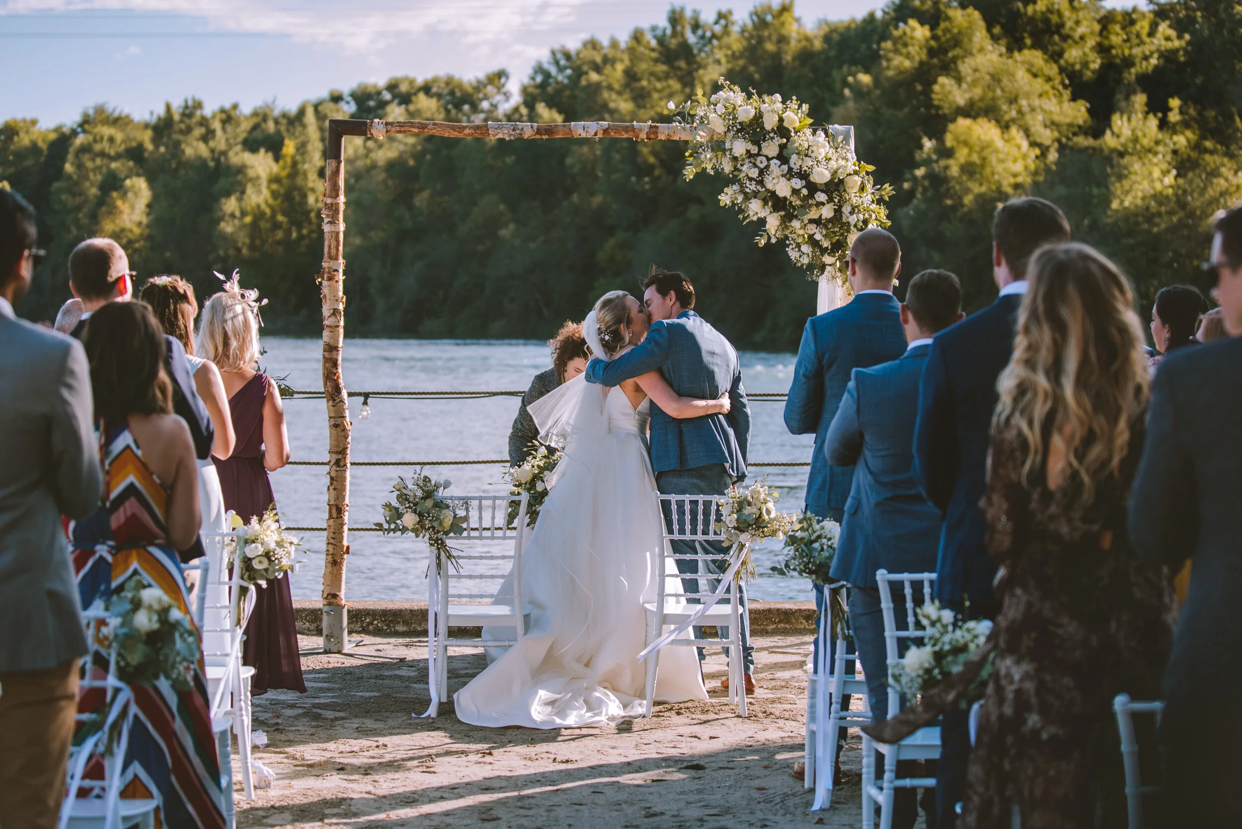 A couple shares a kiss at a wedding ceremony on a riverside, with guests watching and a floral arch behind them.Lake Como Wedding Photographer - Mark Shaw Photography 