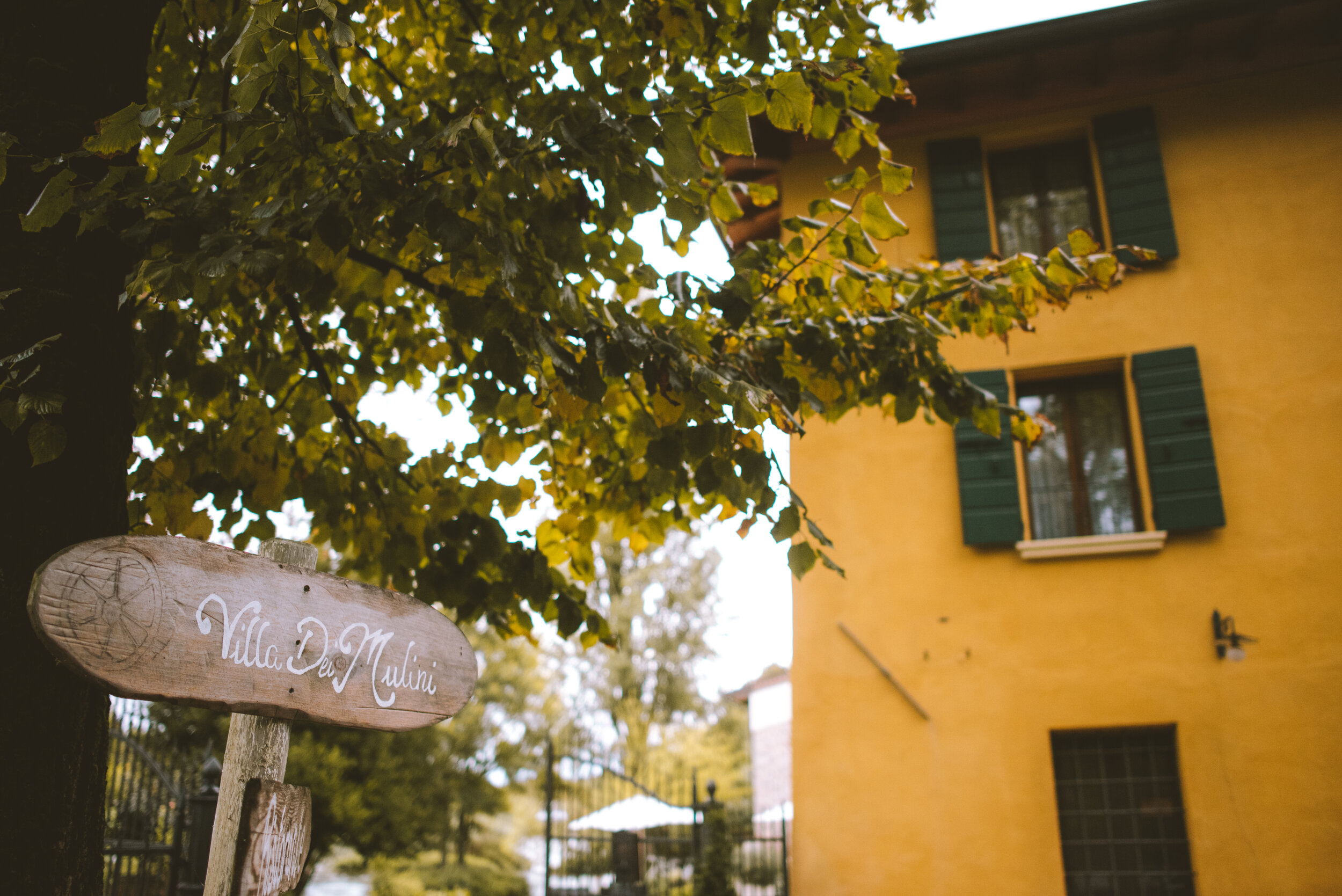 Wooden sign with 'Villa Dei Mulini' written on it, attached to a tree with yellowing leaves in front of a yellow building with green shutters.