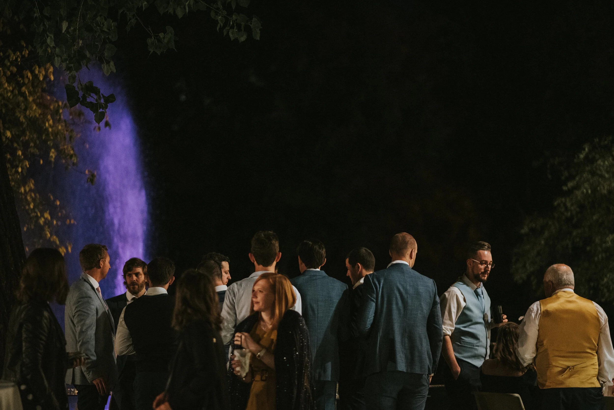 Group of people socializing outdoors at night, with a purple-lit waterfall in the background.