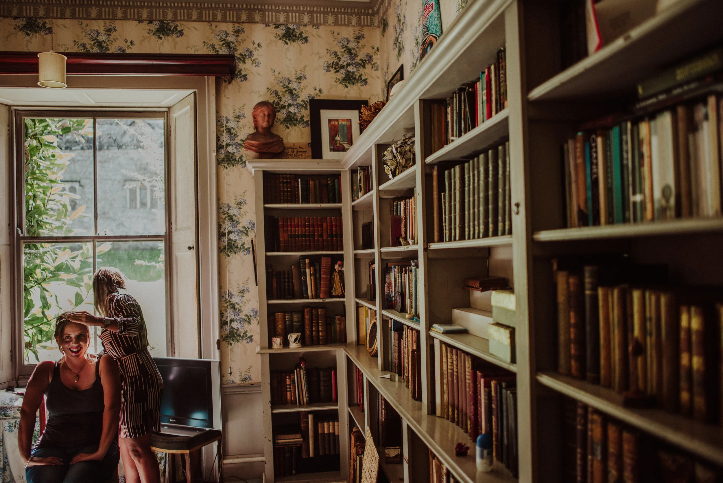 Two women, one seated and smiling and the other standing and combing her hair, in a cozy room with tall bookshelves filled with books, a large window letting in natural light, and vintage decor.