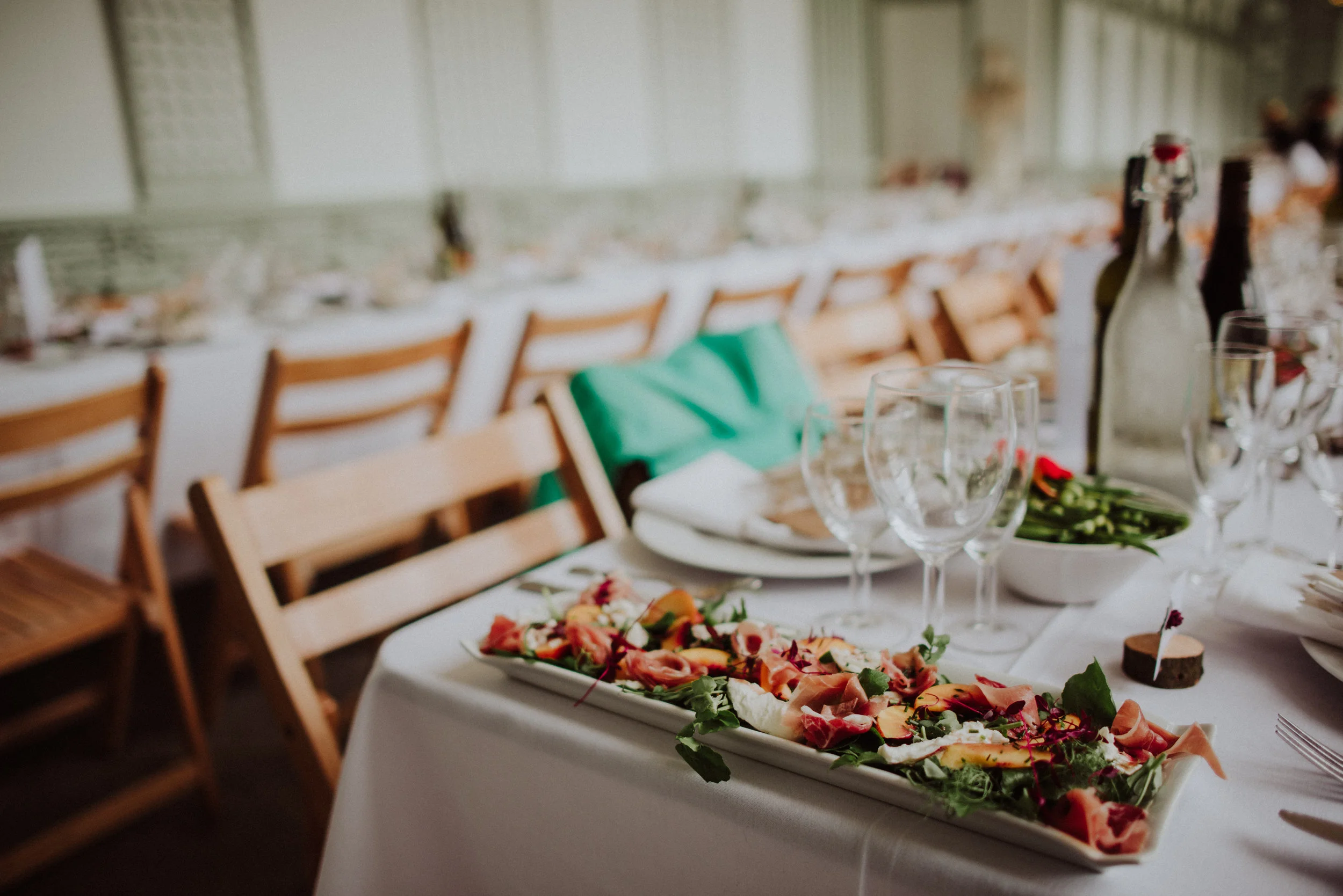 A formal dining table set for a special event with a platter of sliced cured meats and vegetables, wine glasses, bottles, and a green napkin, in a decorated banquet hall.