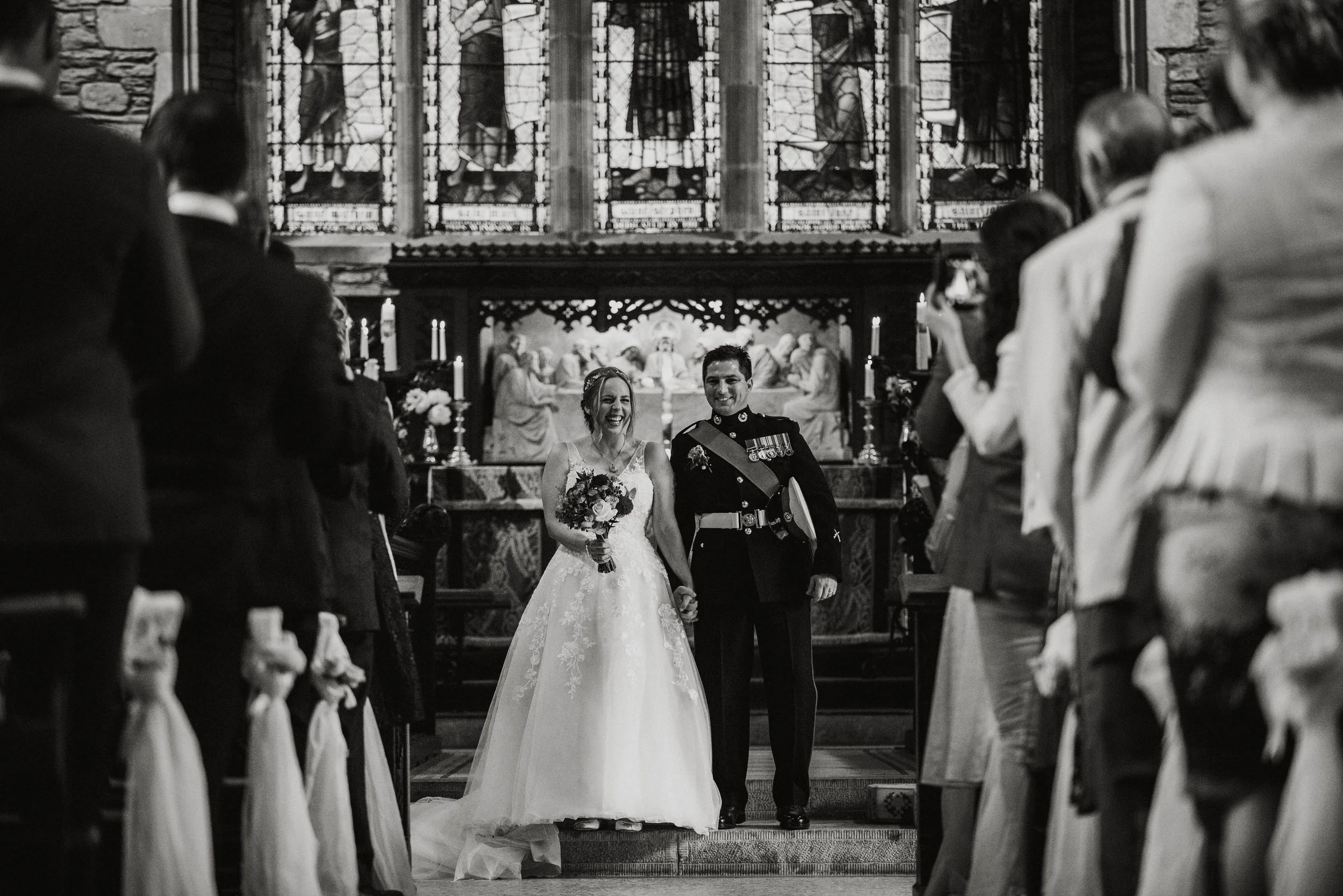 A bride and groom holding hands and walking down the aisle of a church after their wedding ceremony, surrounded by friends and family. The bride is in a white wedding gown holding a bouquet of flowers, and the groom is in a military uniform. The chur