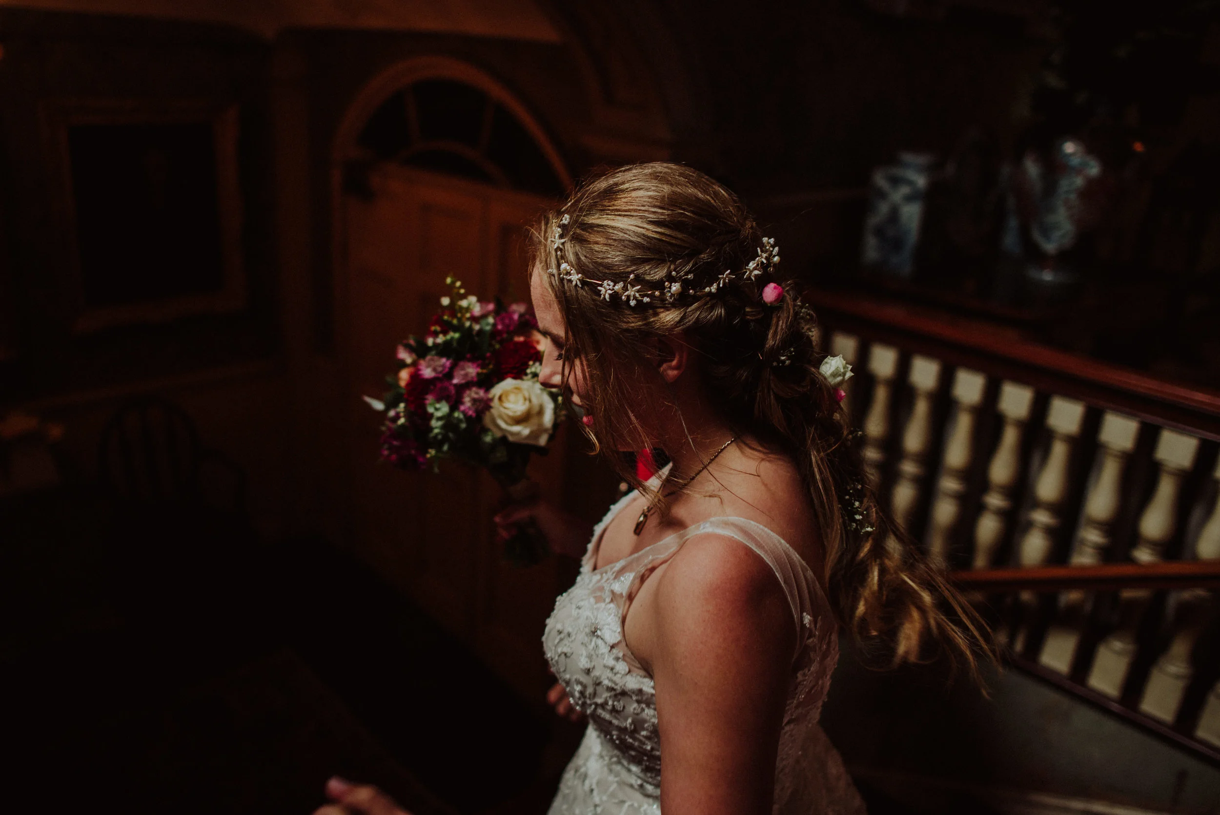 A bride with long wavy hair wearing a lace wedding dress and floral hair accessories, holding a bouquet of flowers, standing on a staircase in a dimly lit wooden interior.