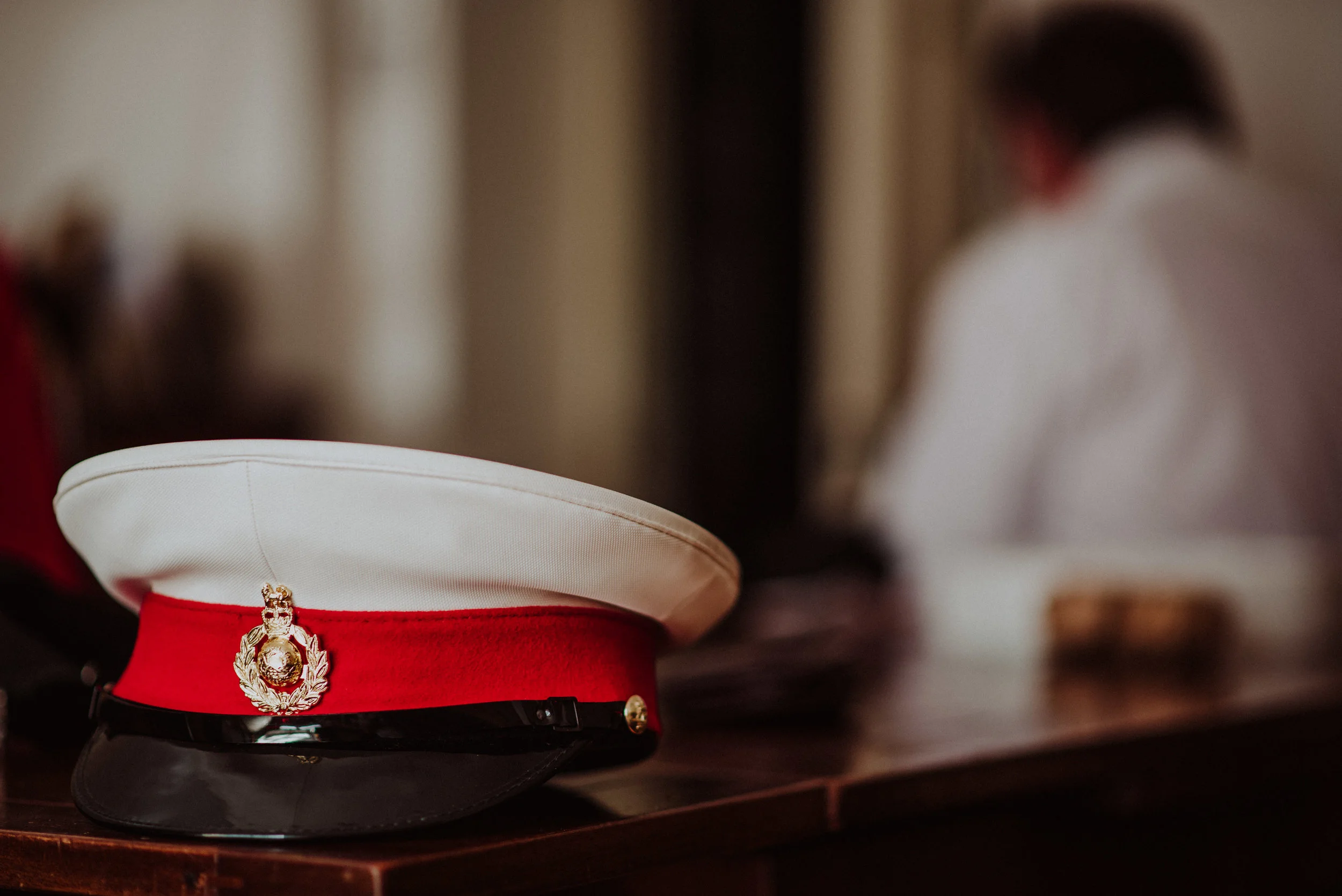 Close-up of a police or military cap with a white top, red band, black brim, and a gold emblem, placed on a wooden surface. Blurred background with a person in a white uniform.