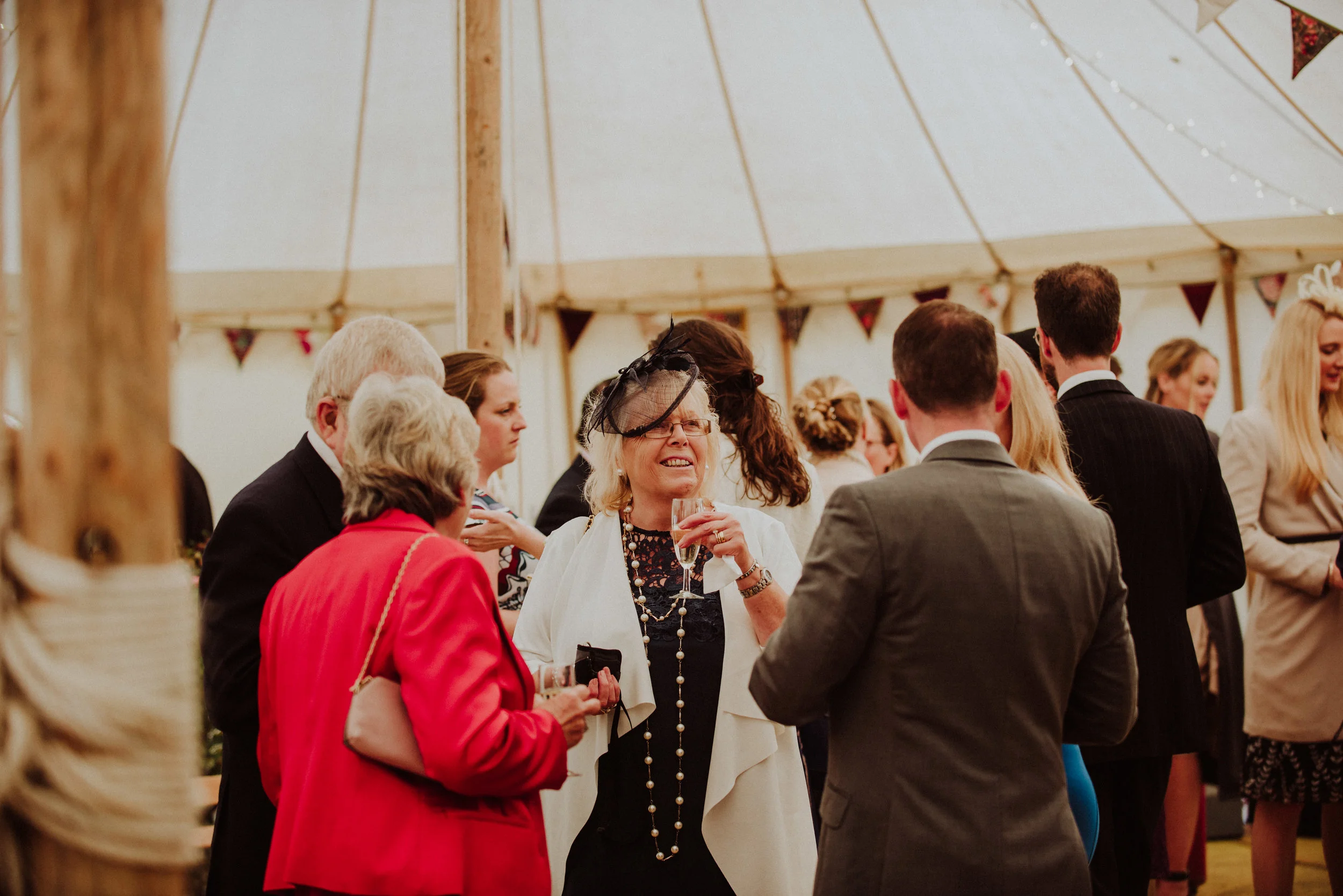 Group of people at a social event inside a large tent, with some engaged in conversation, and a woman in the center holding a glass of drink and smiling.