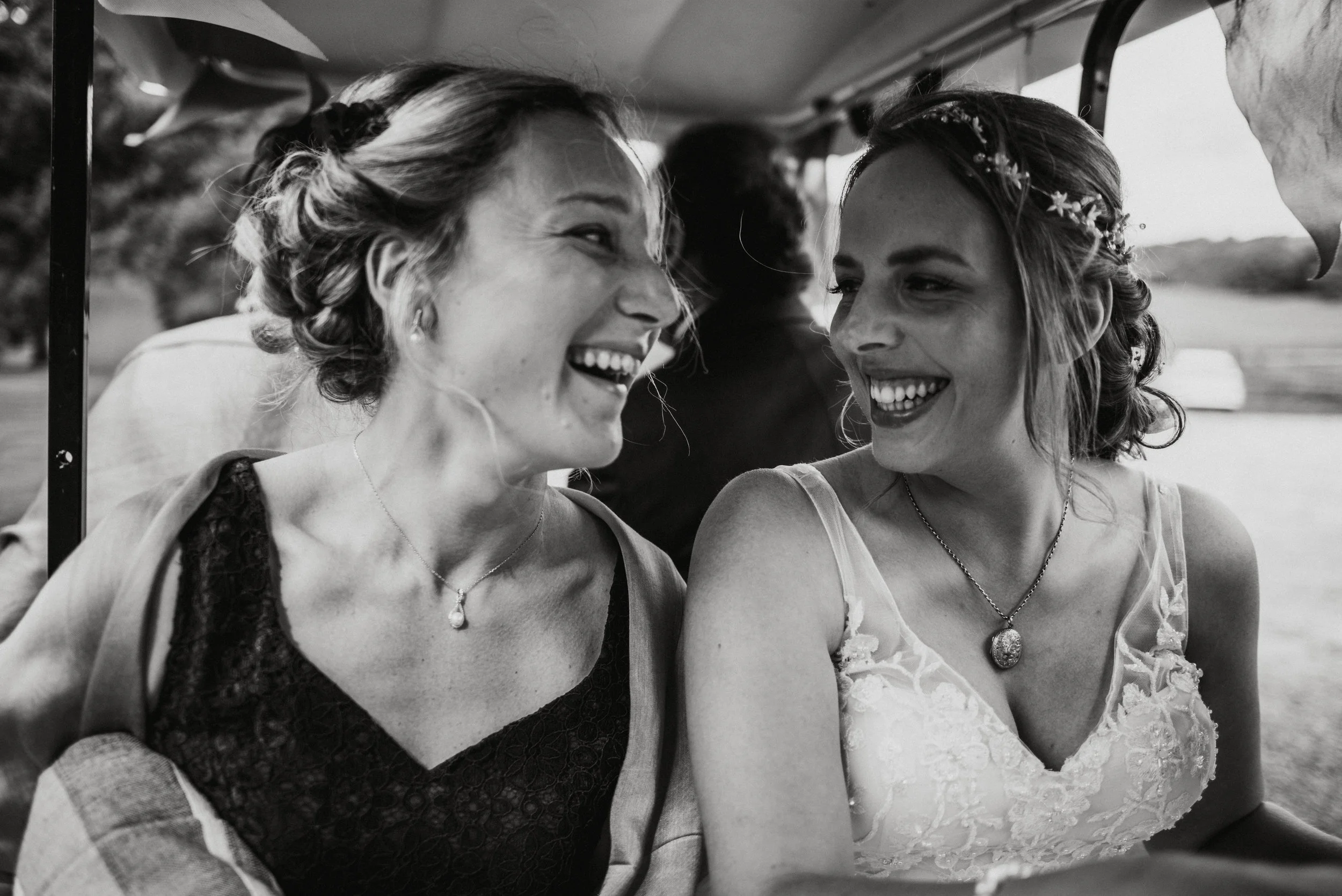 Two women on a golf cart, smiling and looking at each other, in an outdoor setting with trees and mountains in the background, one wearing a wedding dress and the other a dark lace dress.