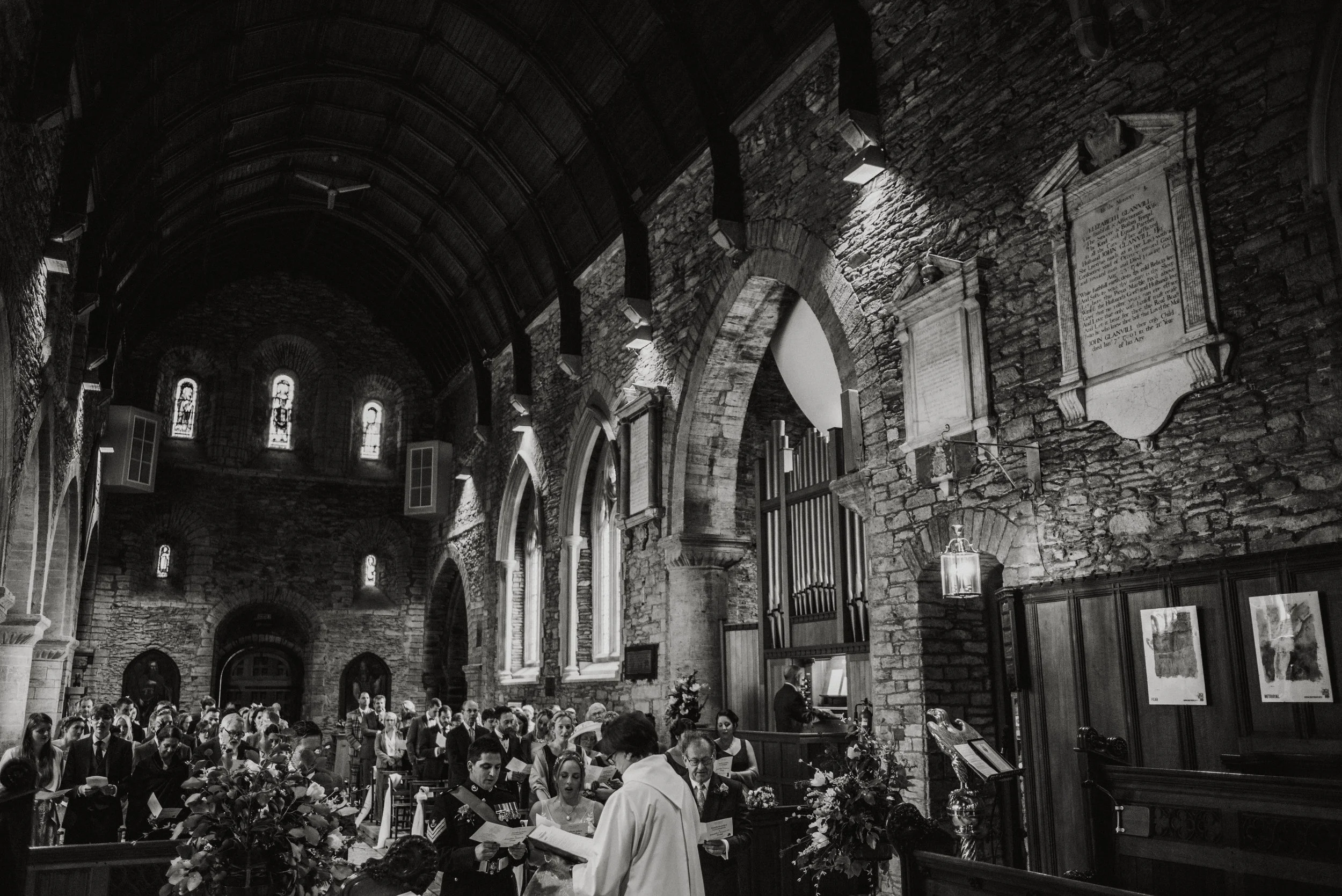 A black and white photo of a wedding ceremony inside a stone church with arched windows and large religious plaques on the walls. The congregation is gathered, and a priest is officiating the ceremony.
