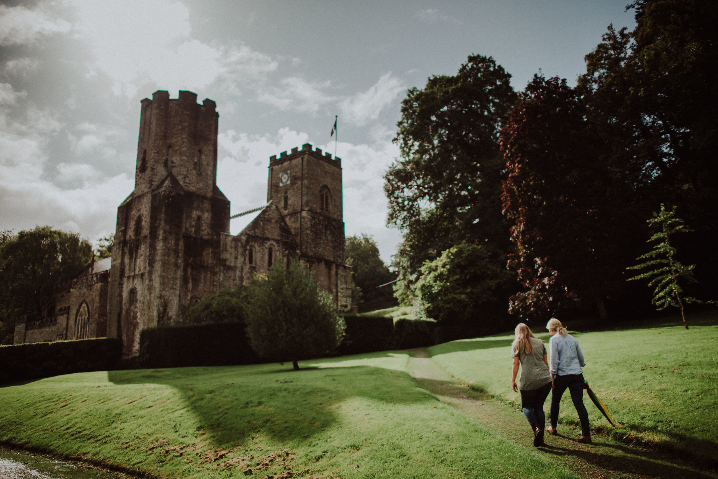 Two women walking on a grassy lawn towards a historic stone castle with towers and battlements, surrounded by trees under a partly cloudy sky.