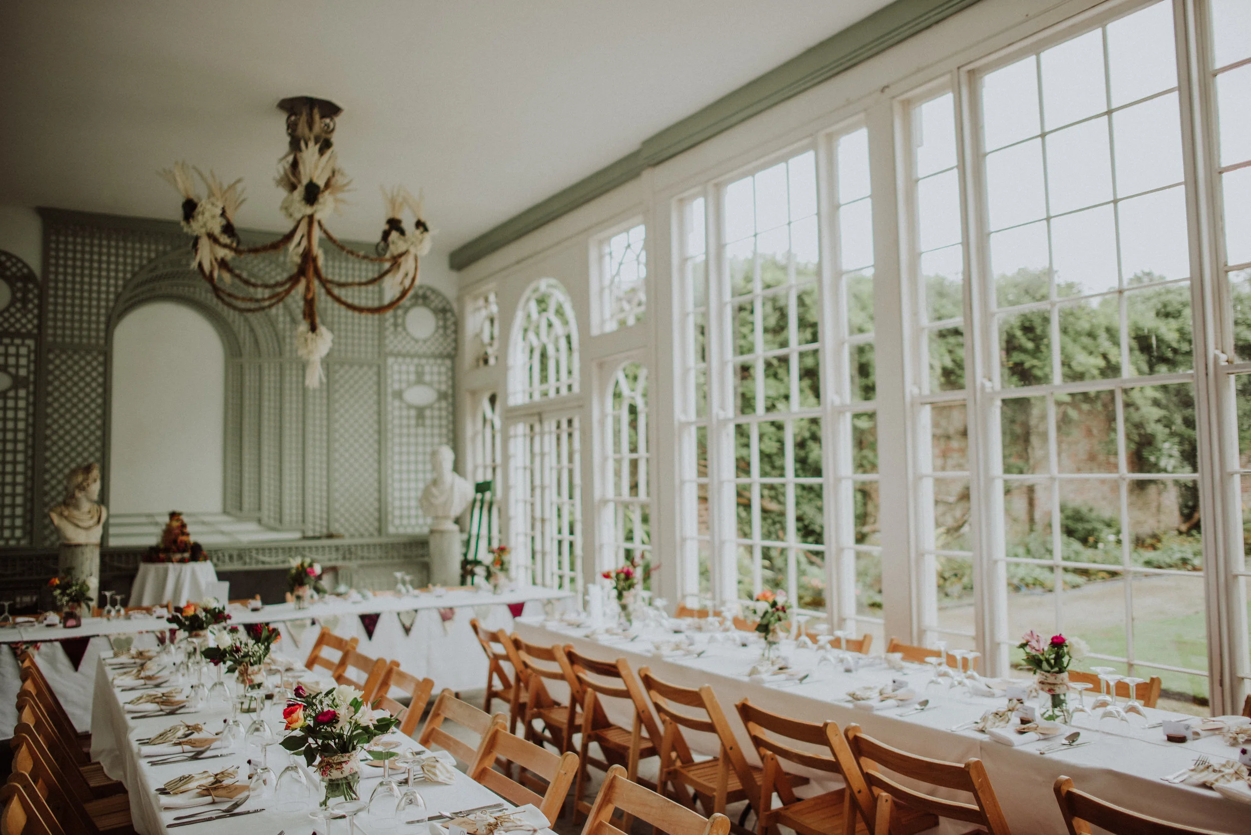 Elegant dining room with large windows, long white table set with flowers and napkins, wooden chairs, bust sculptures, and a chandelier.