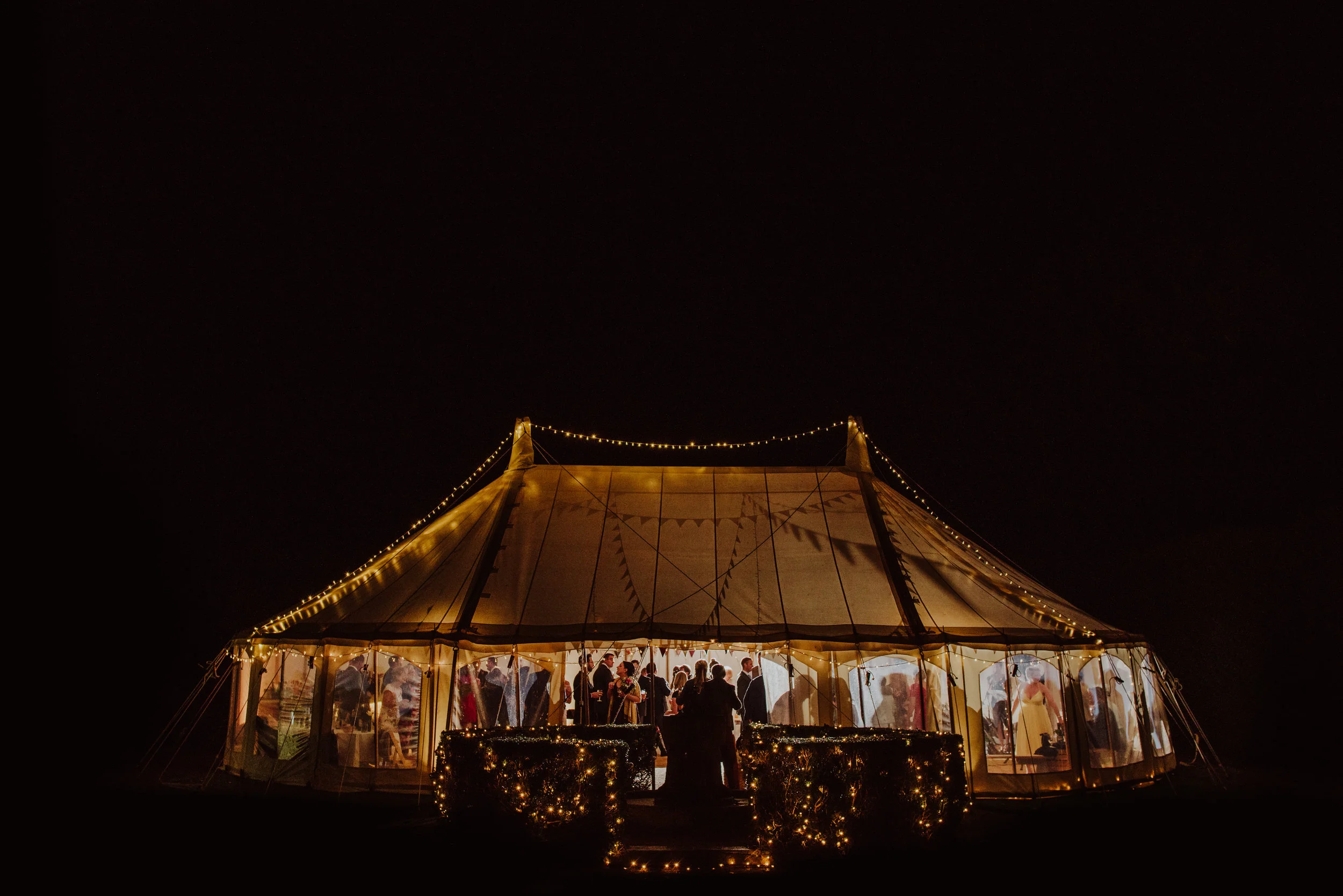 Nighttime celebration inside a large lit-up tent with people gathered, seen through transparent sides, and decorative string lights around the entrance.